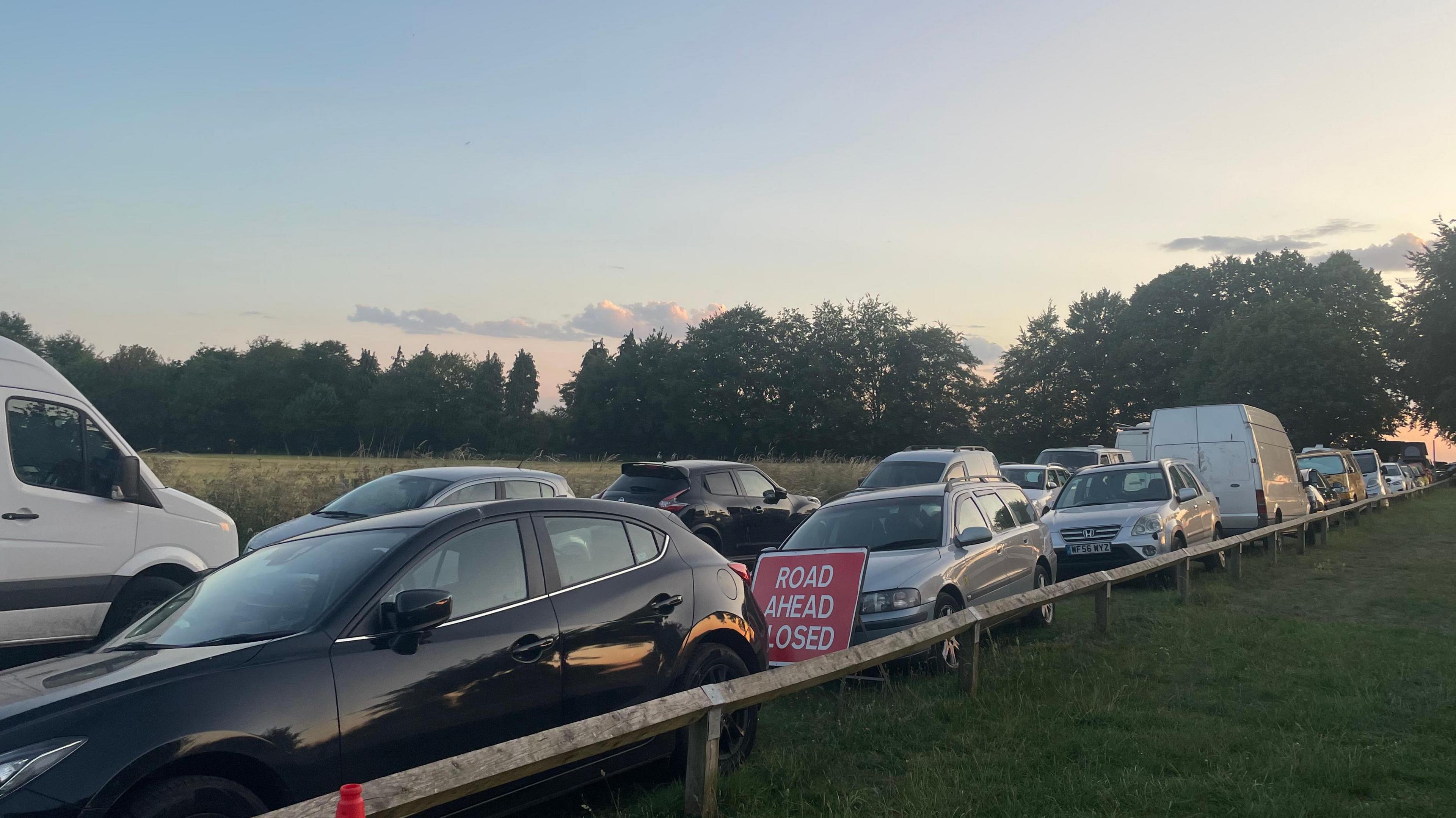 Cars parked close together, on grass verges either side of a small country road.