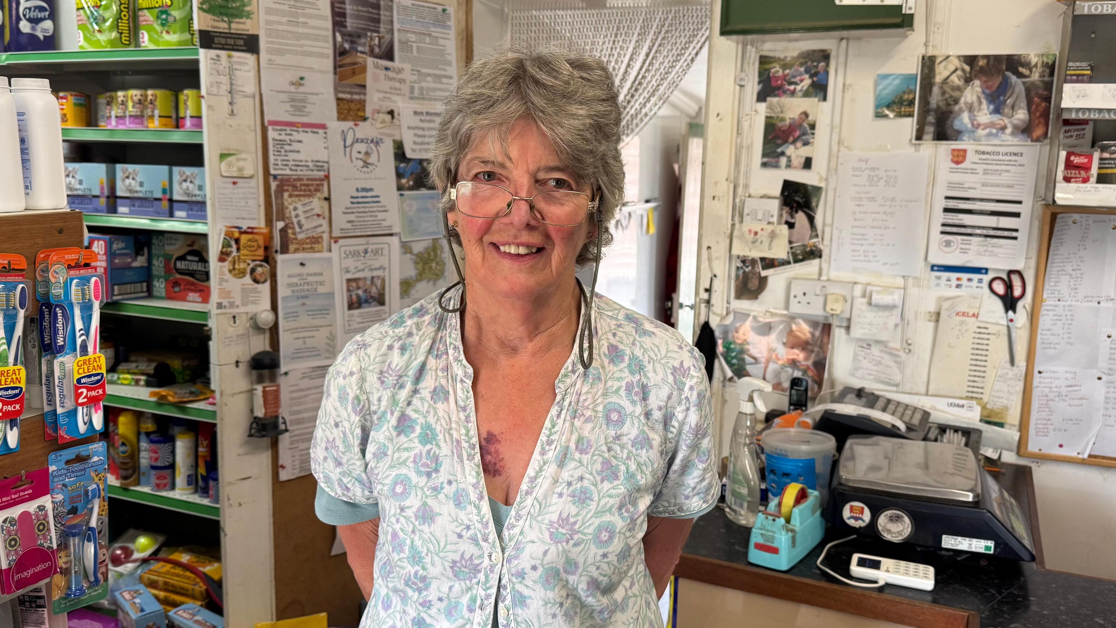 A lady with grey short hair standing in a shop. there are toothbrushes, petfood and a till in the background.