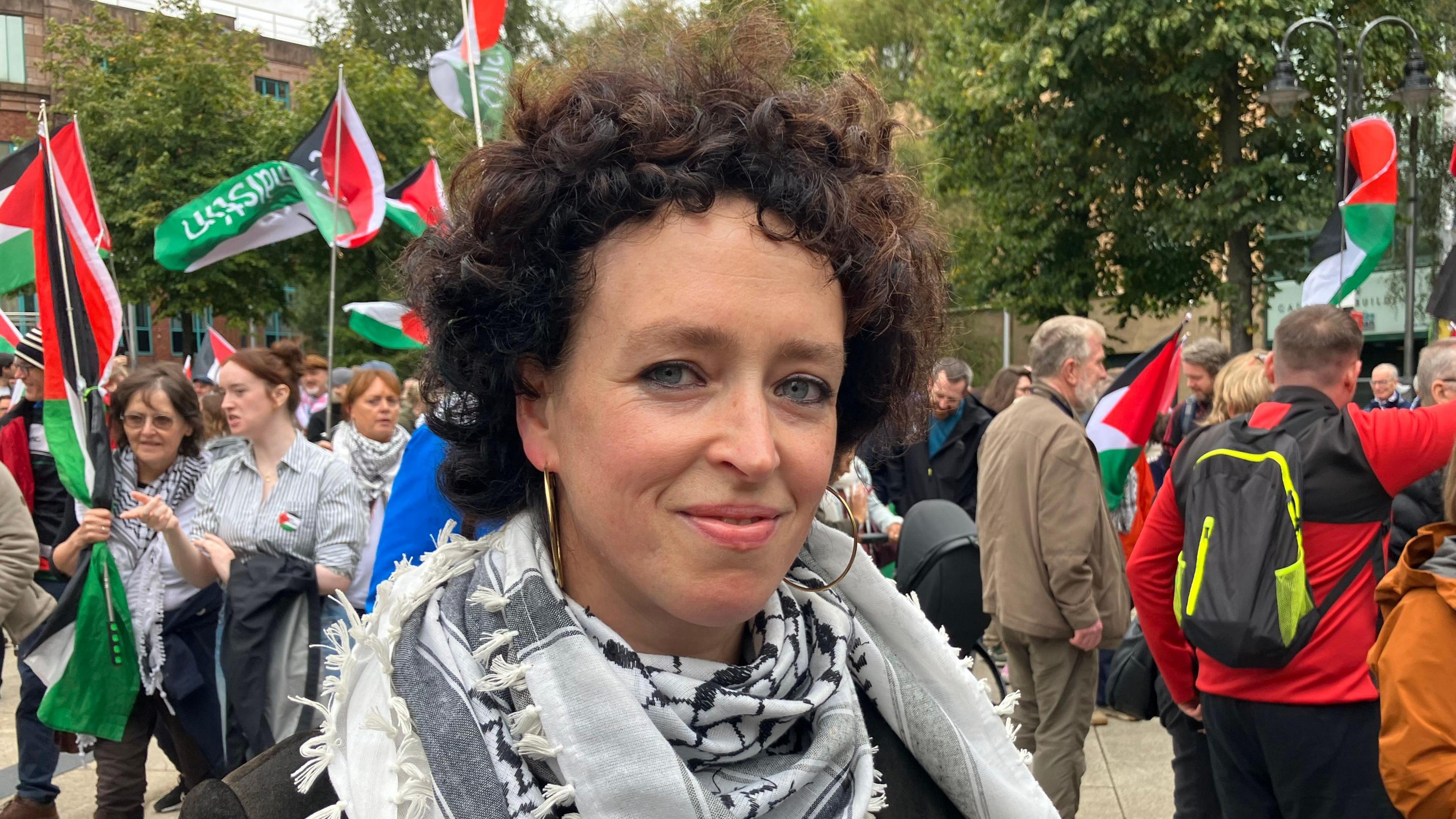 Orla Wallace smiling at the camera during the Belfast protest. She has dark, curly hair and is weaing gold hooped earrings and a black and white scarf. People behind her are carrying Palestinian flags.
