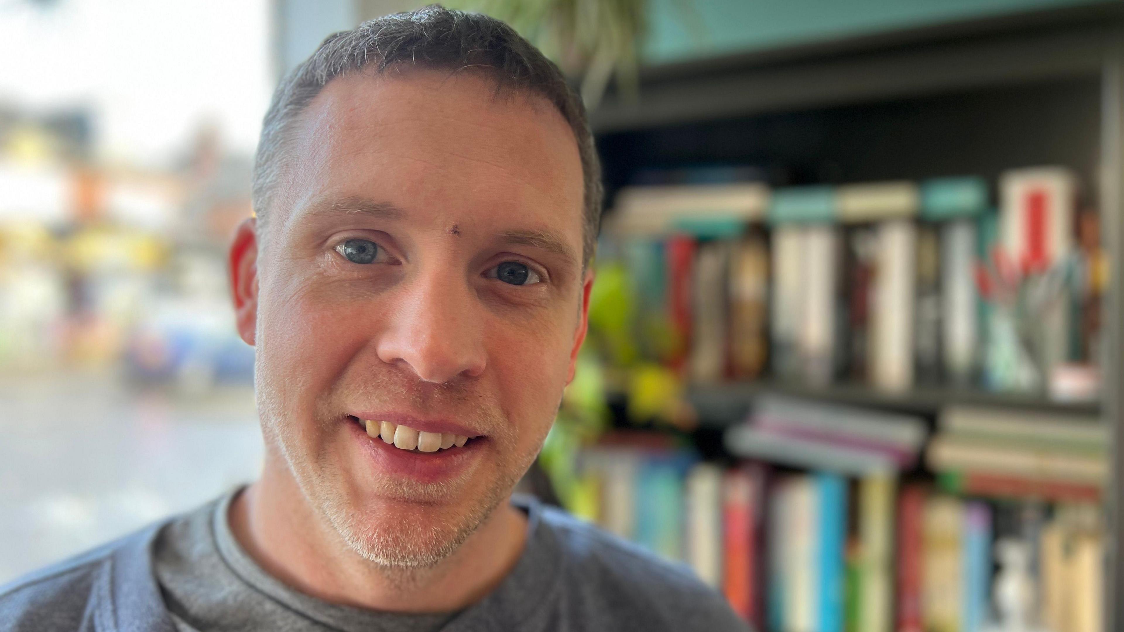 A man with short hair and stubble smiling at the camera, behind him is a book shelf stacked with paperbacks.