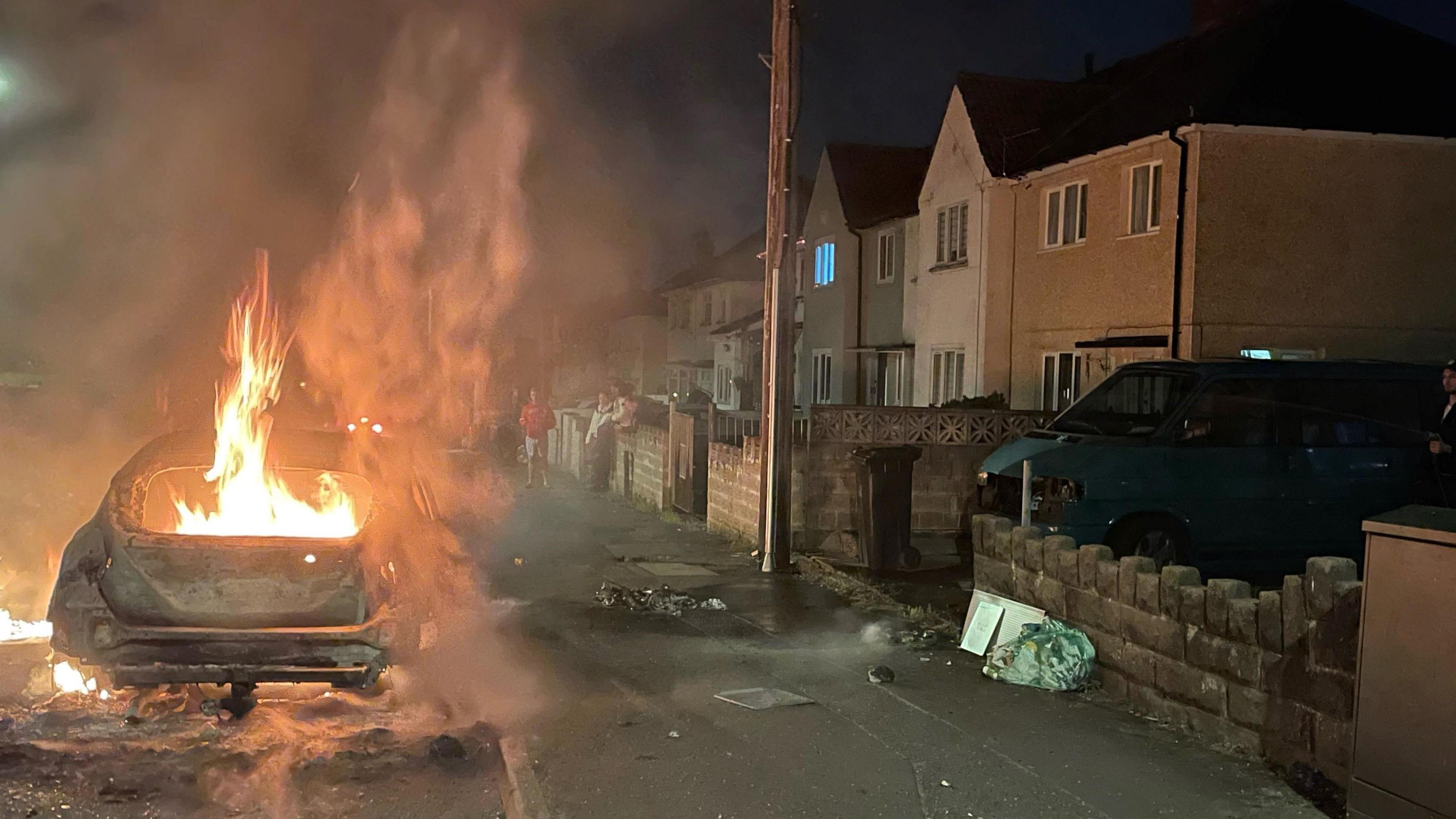 A car which is on fire. It is parked next to a pavement with a row of house on the rigth hand side.