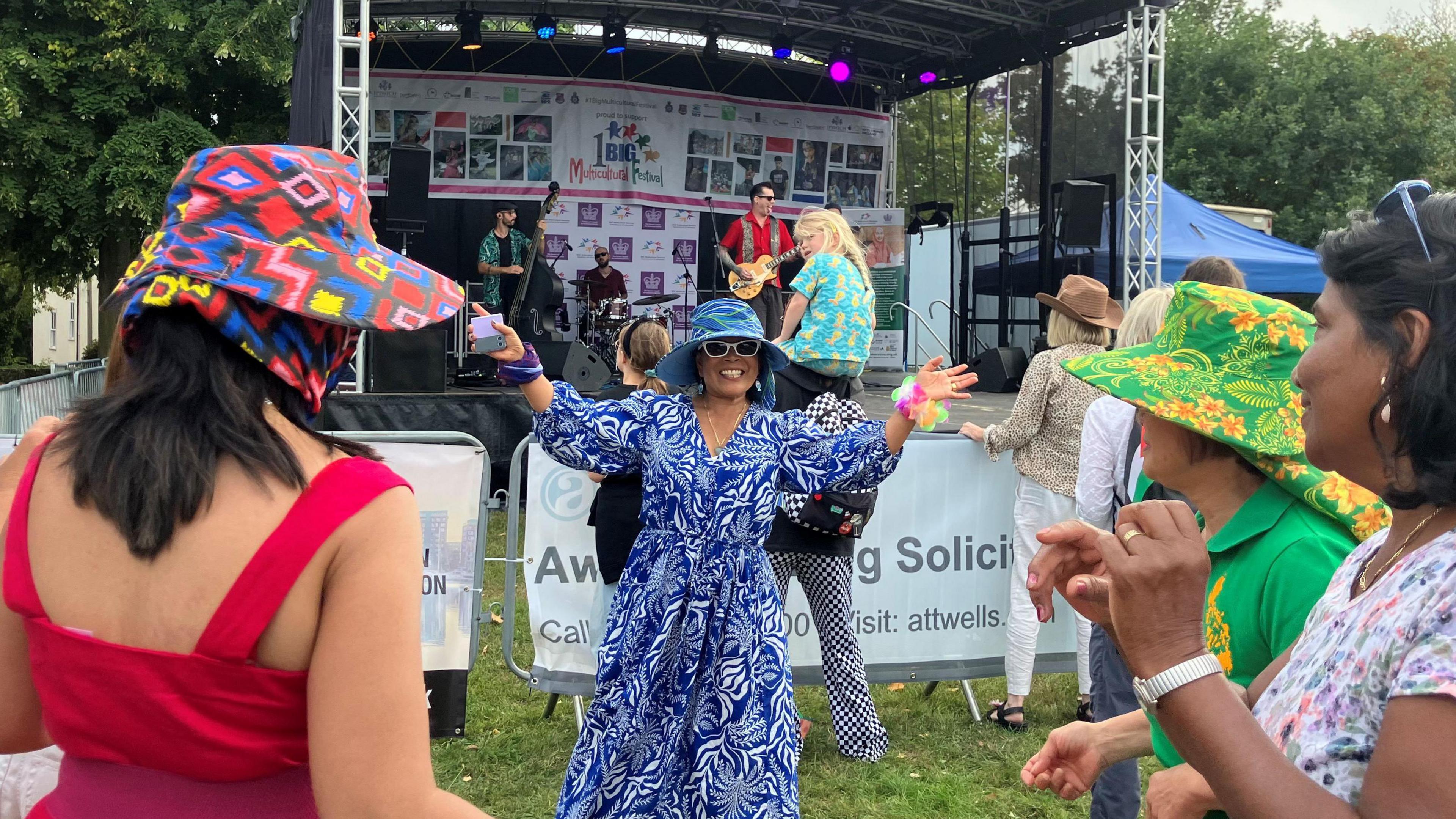 Several women in bight outfits dance in front of a festival stage.