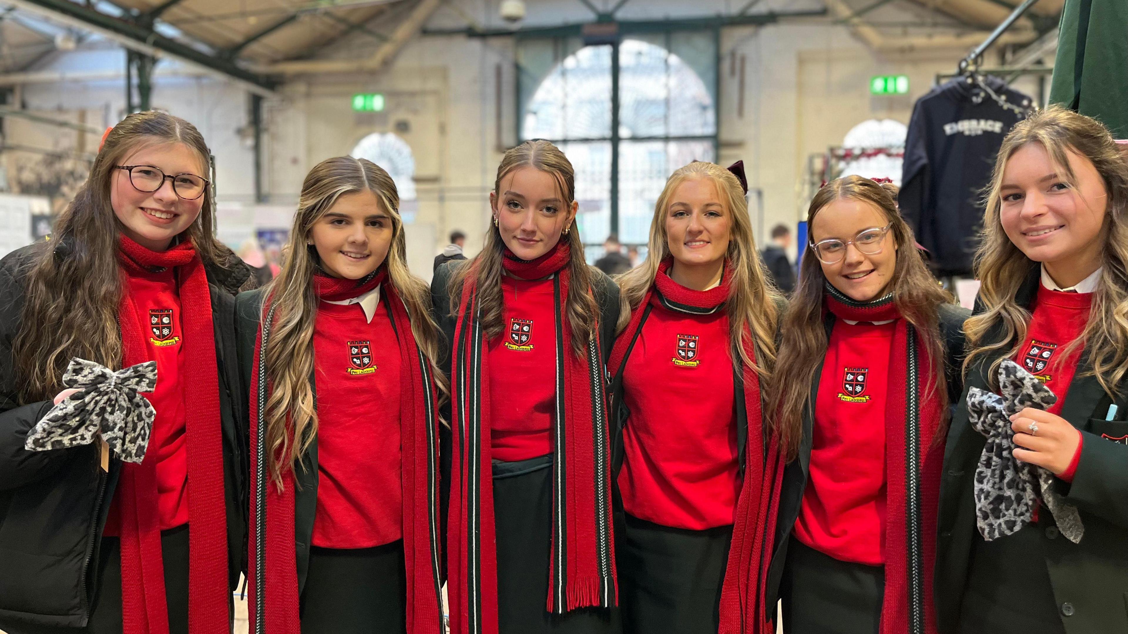 Six girls wearing red jumpers and dark green skirts and blazers. They have linked arms and are standing in the middle of St George's Market. All apart from the girl on the far right are wearing long red scarves with green and white detail.