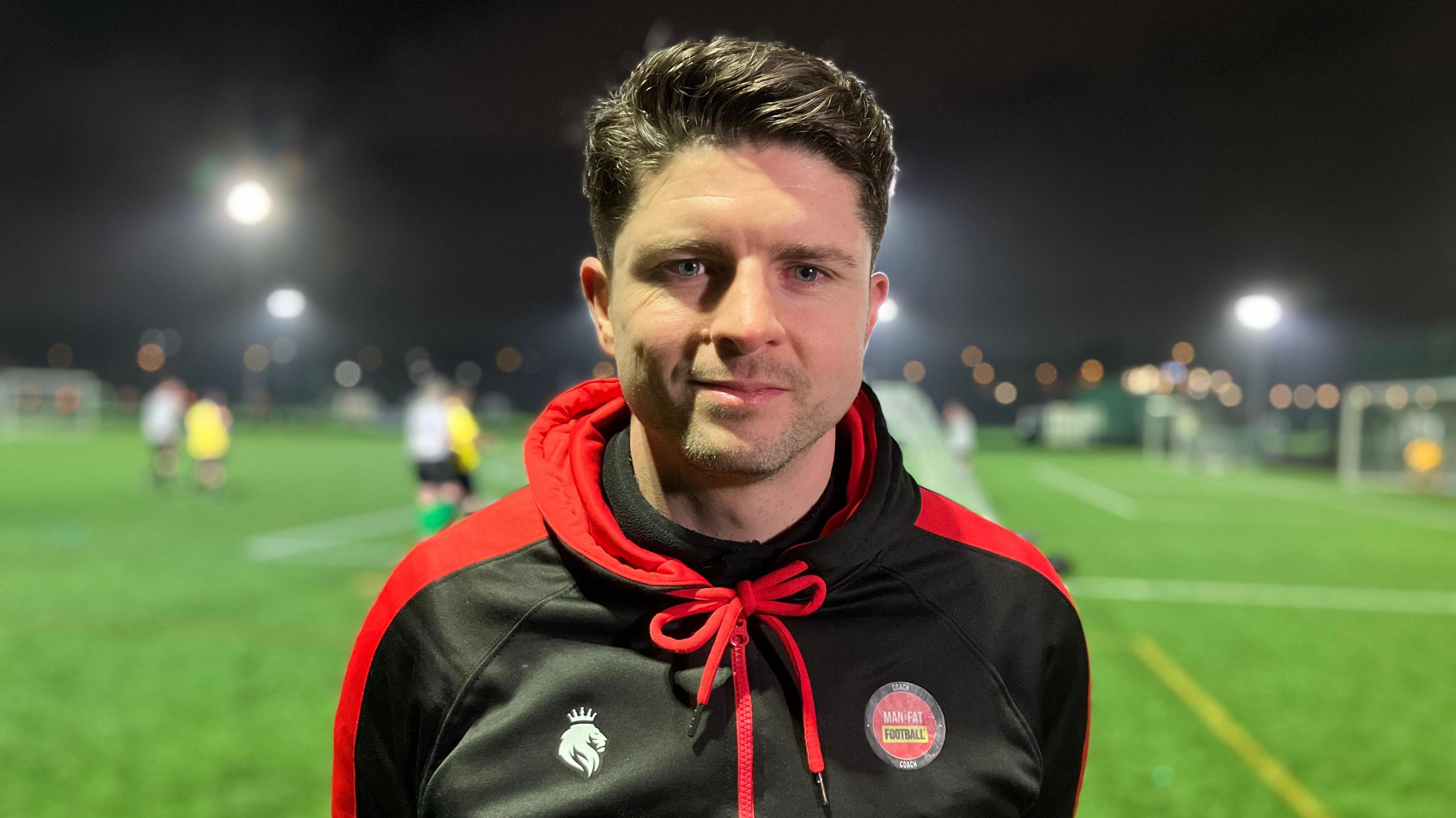 Coach Steve James looks into the camera while a football match happens behind him