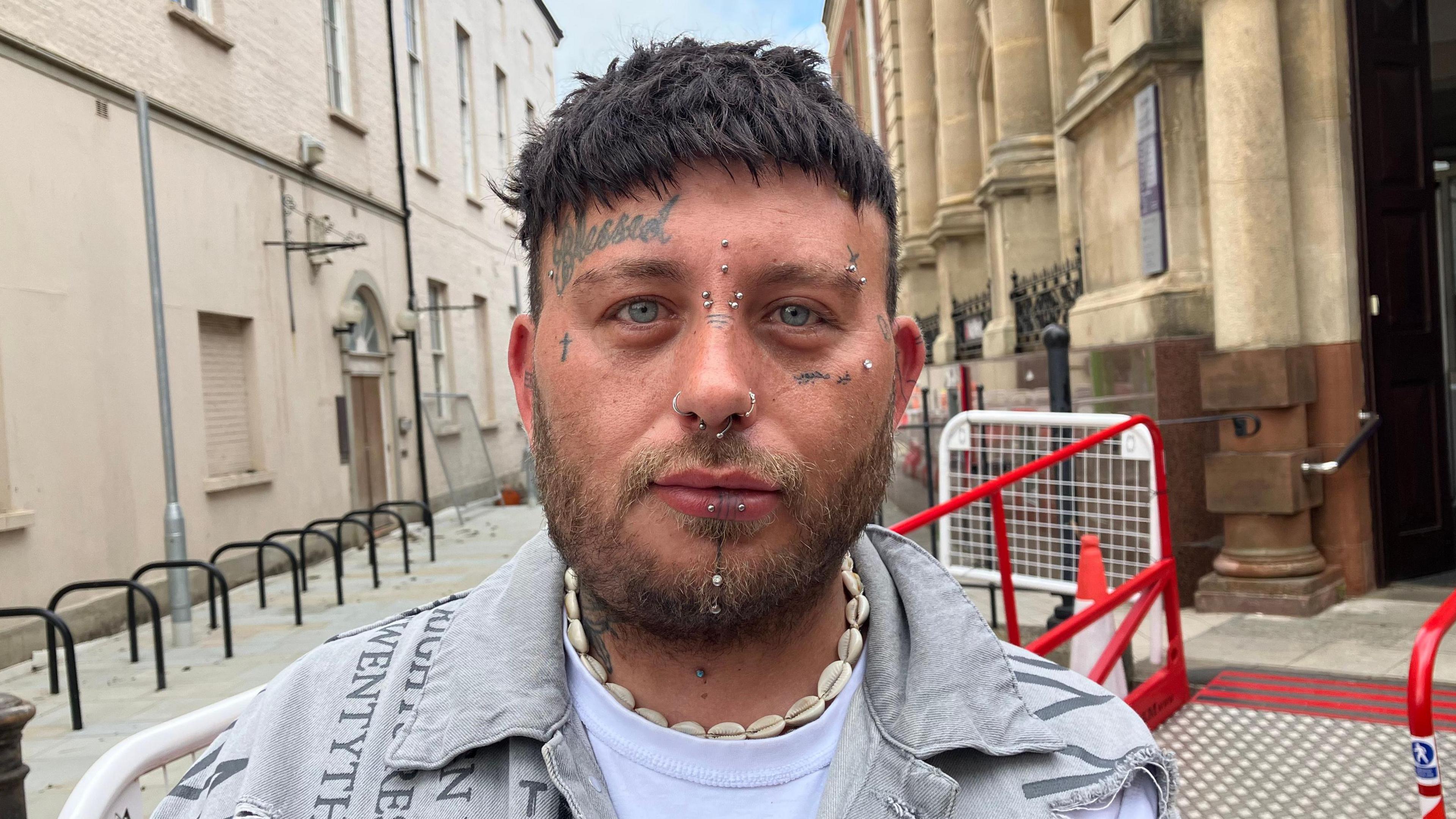Headshot image of a man with facial tattoos and facial piercings, wearing a denim jacket and a shell necklace standing on a street