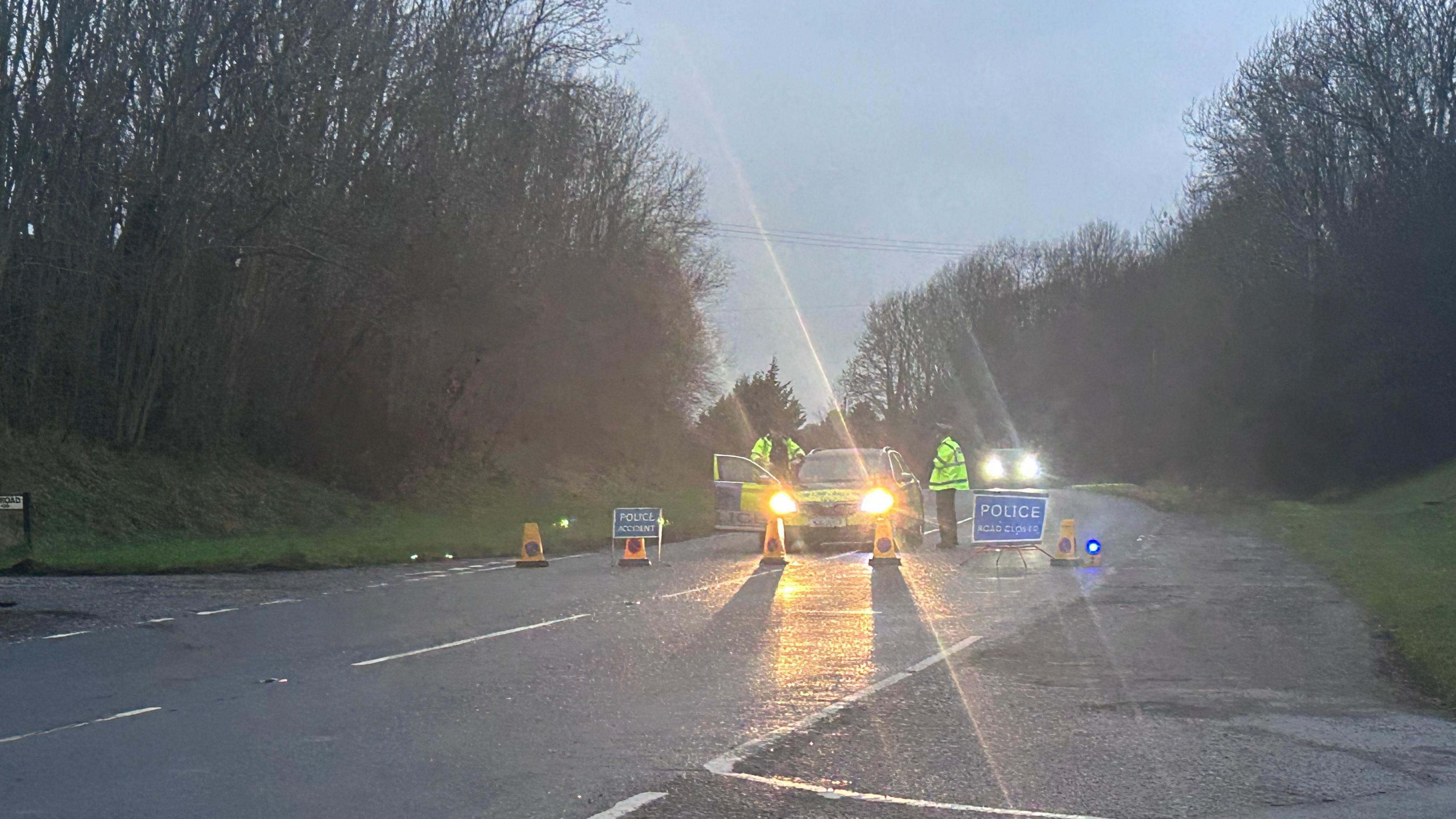 A police car with its headlights on, blocking off the road. Two police officers can be seen wearing high vis yellow jackets. Yellow cones and a police warning sign are on the road in front of the car. 
