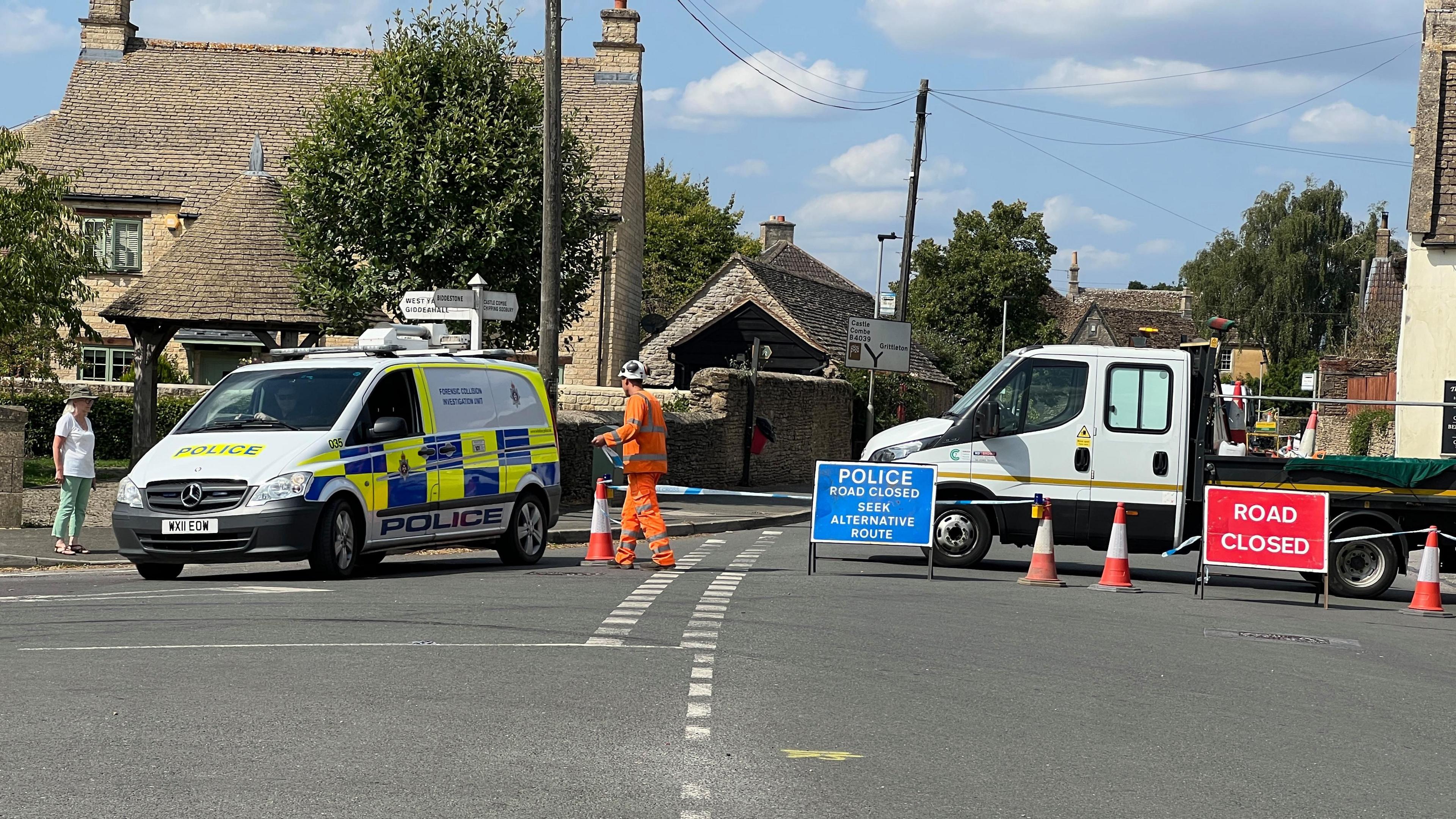 Street in Chippenham with a police van to the left of the frame. On the right, a blue police road closure sign blocks the road, along with a white van. Police tape cordons off the road. In the background there are houses and blue sky. 