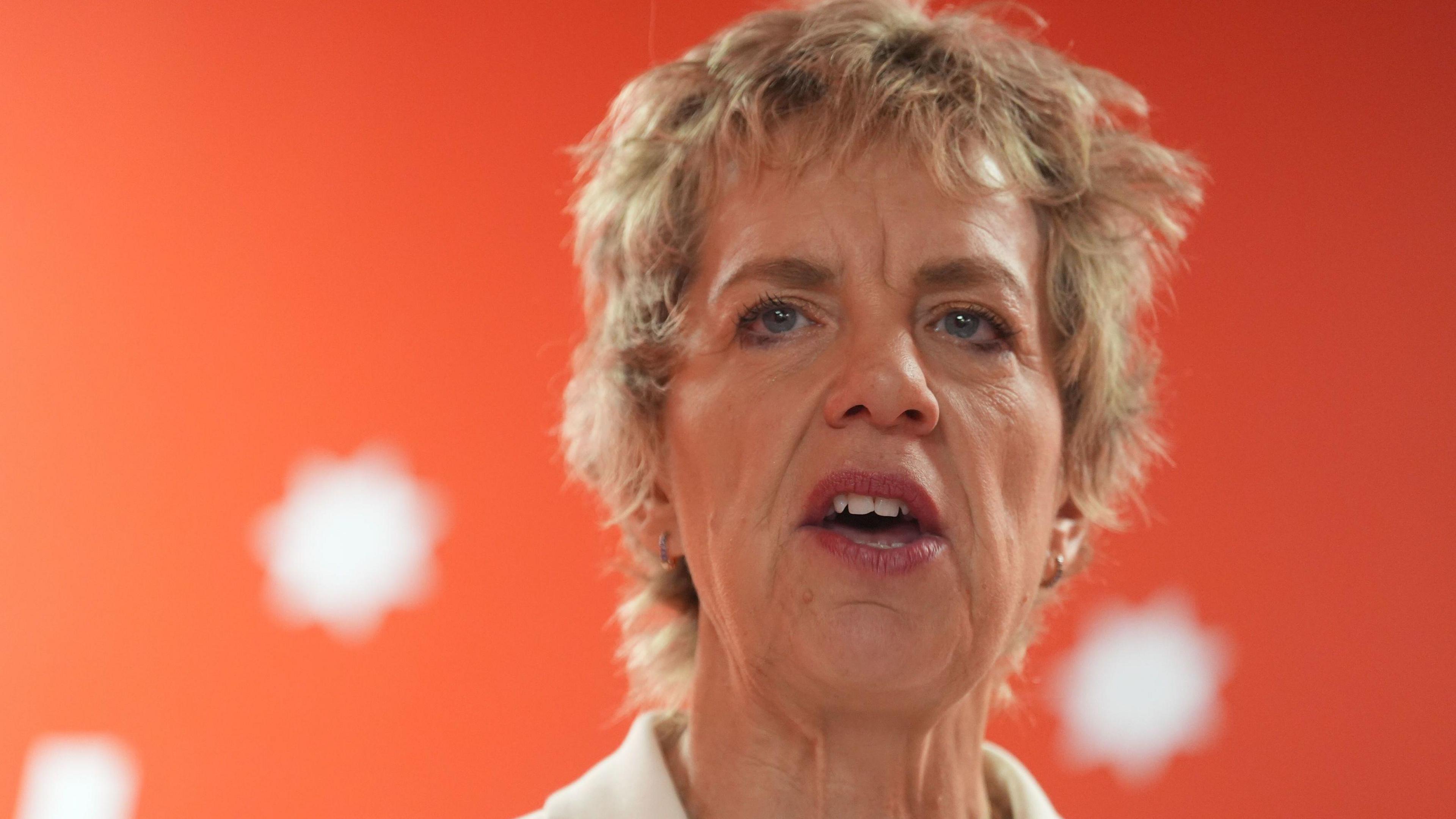 A woman with short blonde hair is pictured talking in front of a red background with white spots. It's a close up but the collar of a white top she is wearing can be seen.