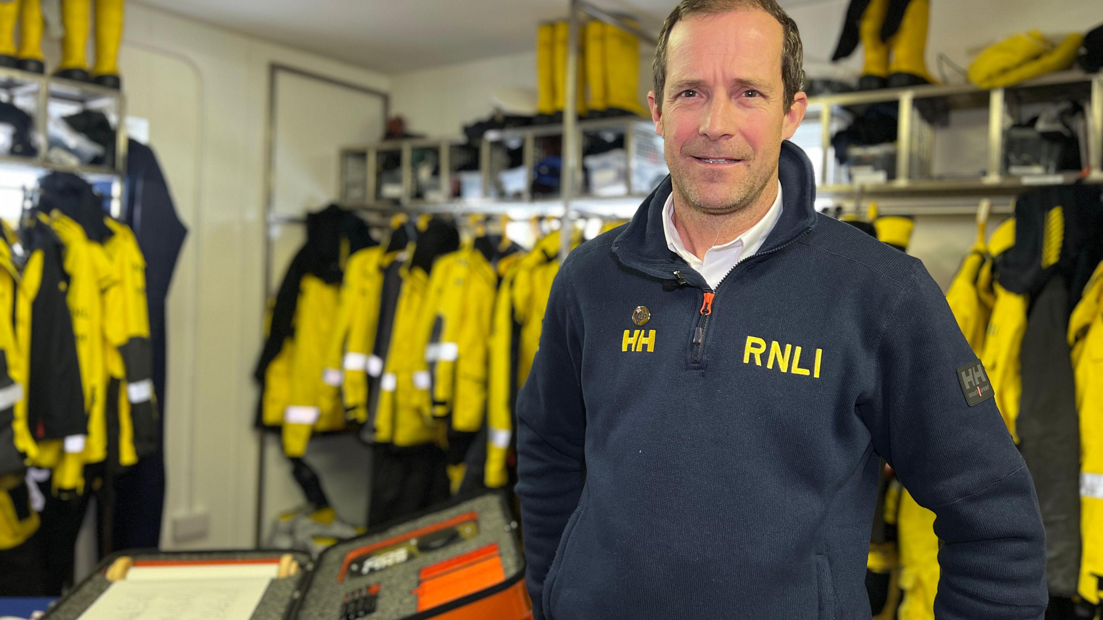 Barclay Harvey, helm of the inshore lifeboat in St Helier, Jersey. He is a middle-aged white man wearing a navy RNLI-branded top. In the background are a series of yellow waterproofs.