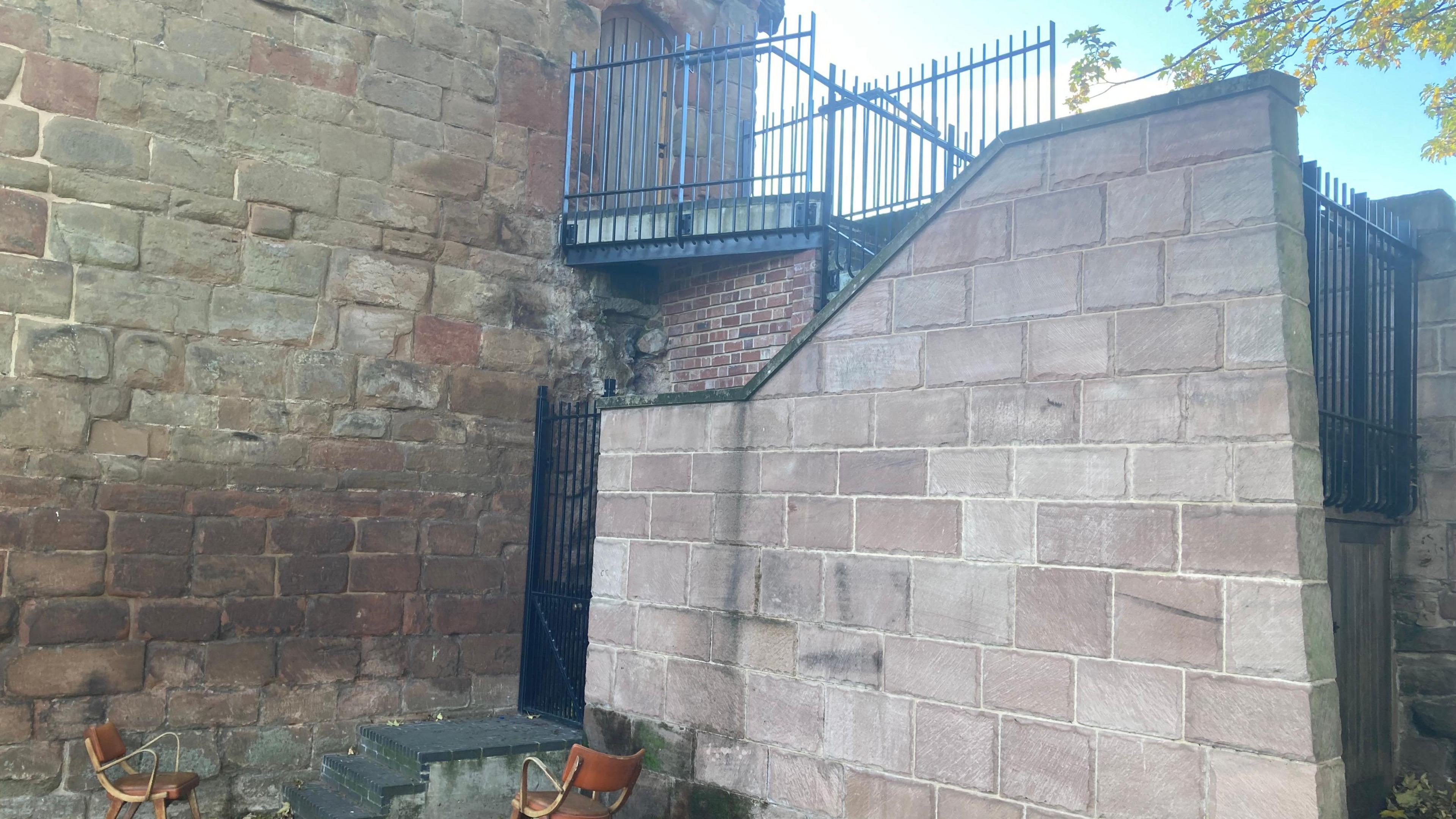 Two wooden chairs have been placed on grass on either side of concrete steps which lead to a black iron gate. This is the entrance to the stairs going up to the Cook Street Gate flat.