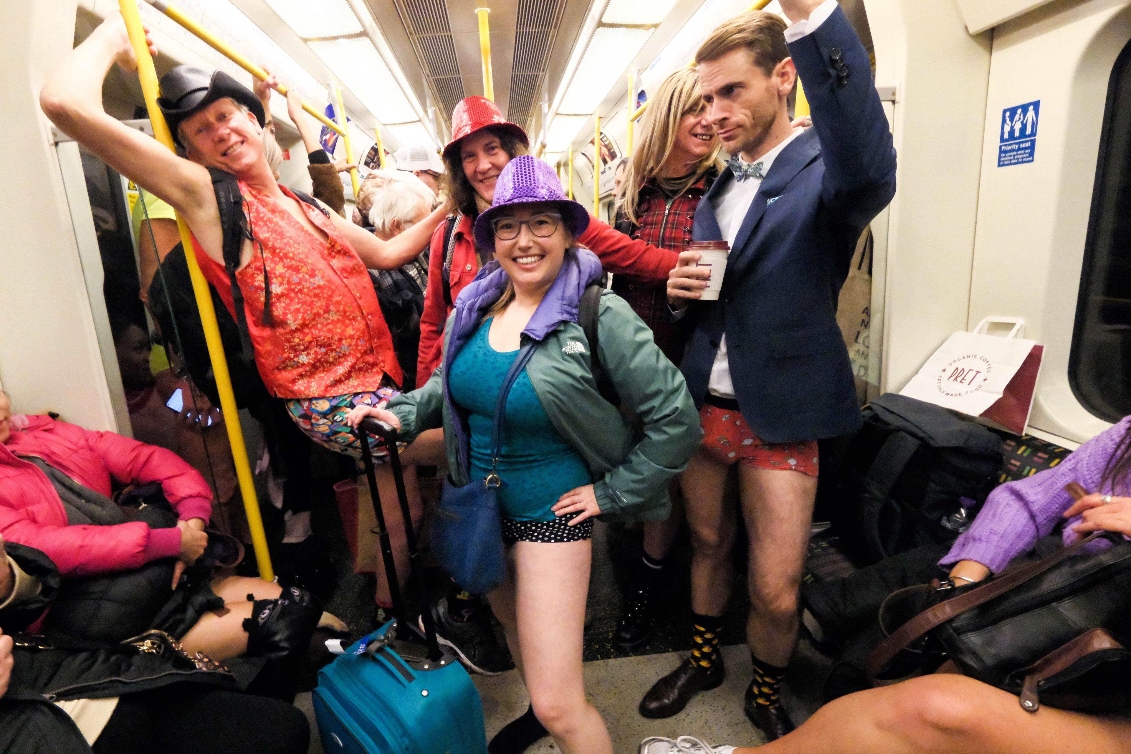 Woman with no trousers wearing purple hat and holding blue suitcase while other passengers smile and a man with no trousers hold a coffee cup