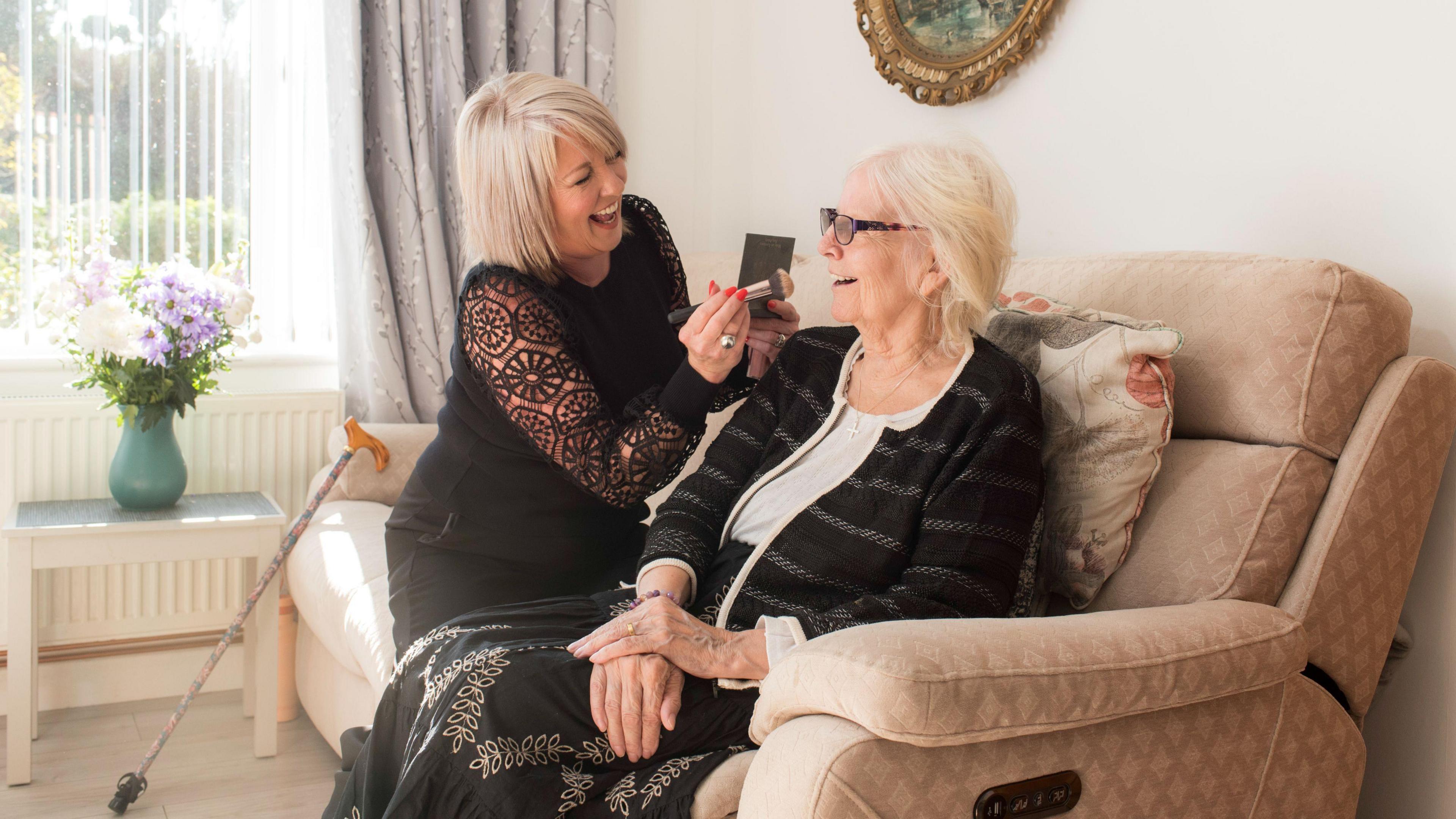 Two women sit on a sofa. One has a make-up brush and seems like she is about to use it on the other. The room is well-lit. They are laughing. 