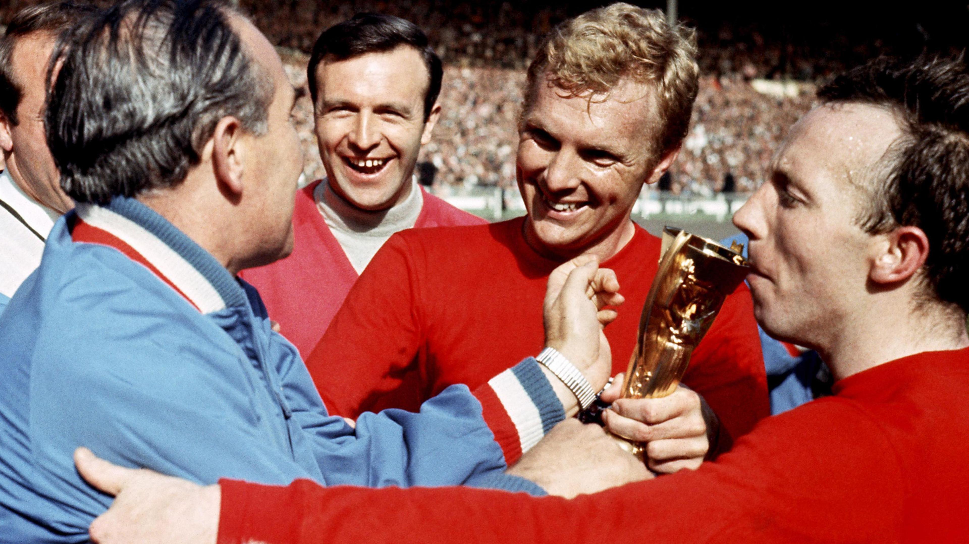 Sir Alf Ramsey in a light blue tracksuit (far left), Jimmy Armfield (centre left) with black hair in light turtle neck shirt and red sweater, Bobby Moore (centre right) with a red England shirt has blonde hair and Nobby Stiles (far right) has balck hair and  red England shirt.
