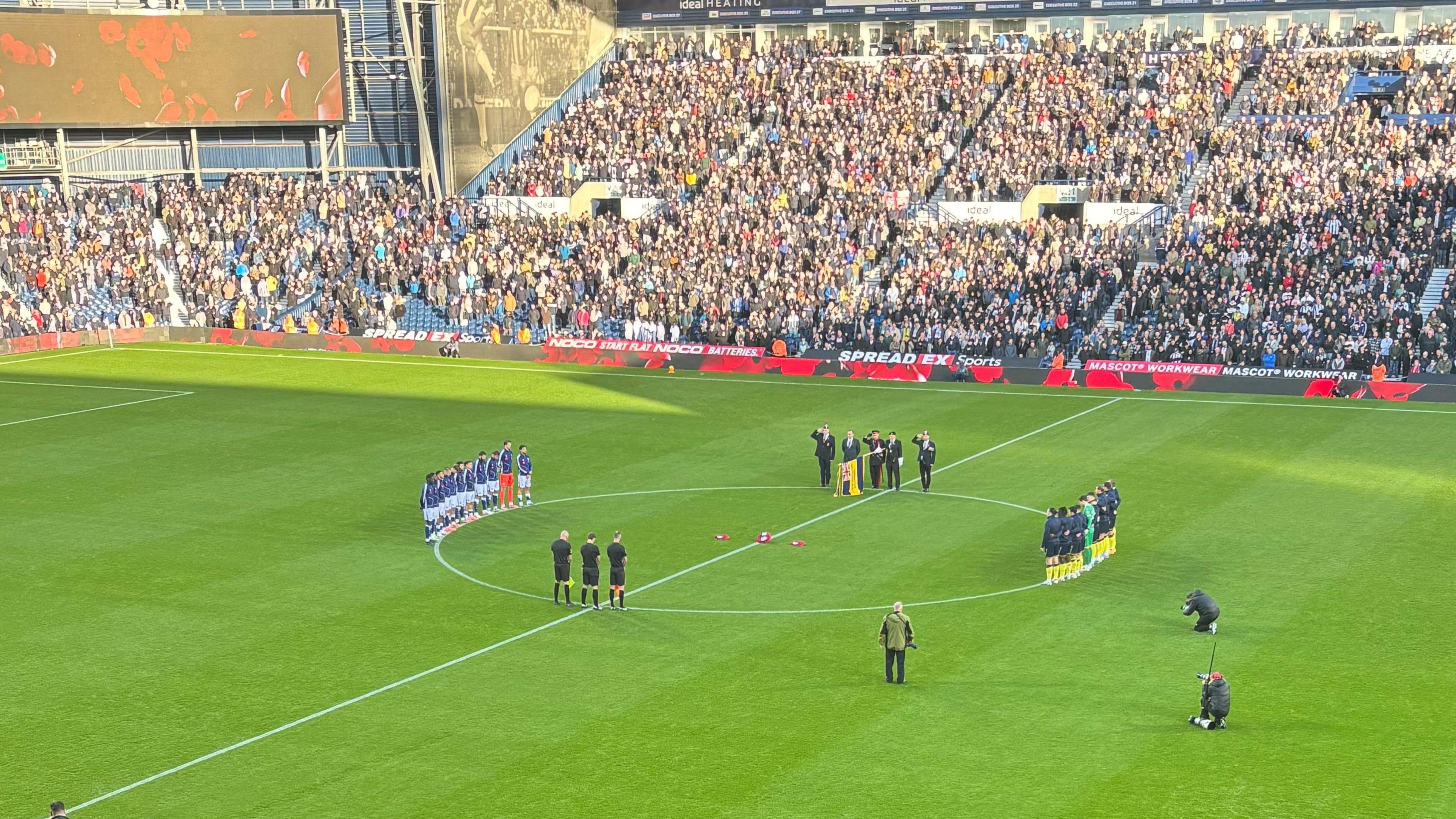 Footballers lined up around the centre circle of a football pitch with people standing in the stands behind them