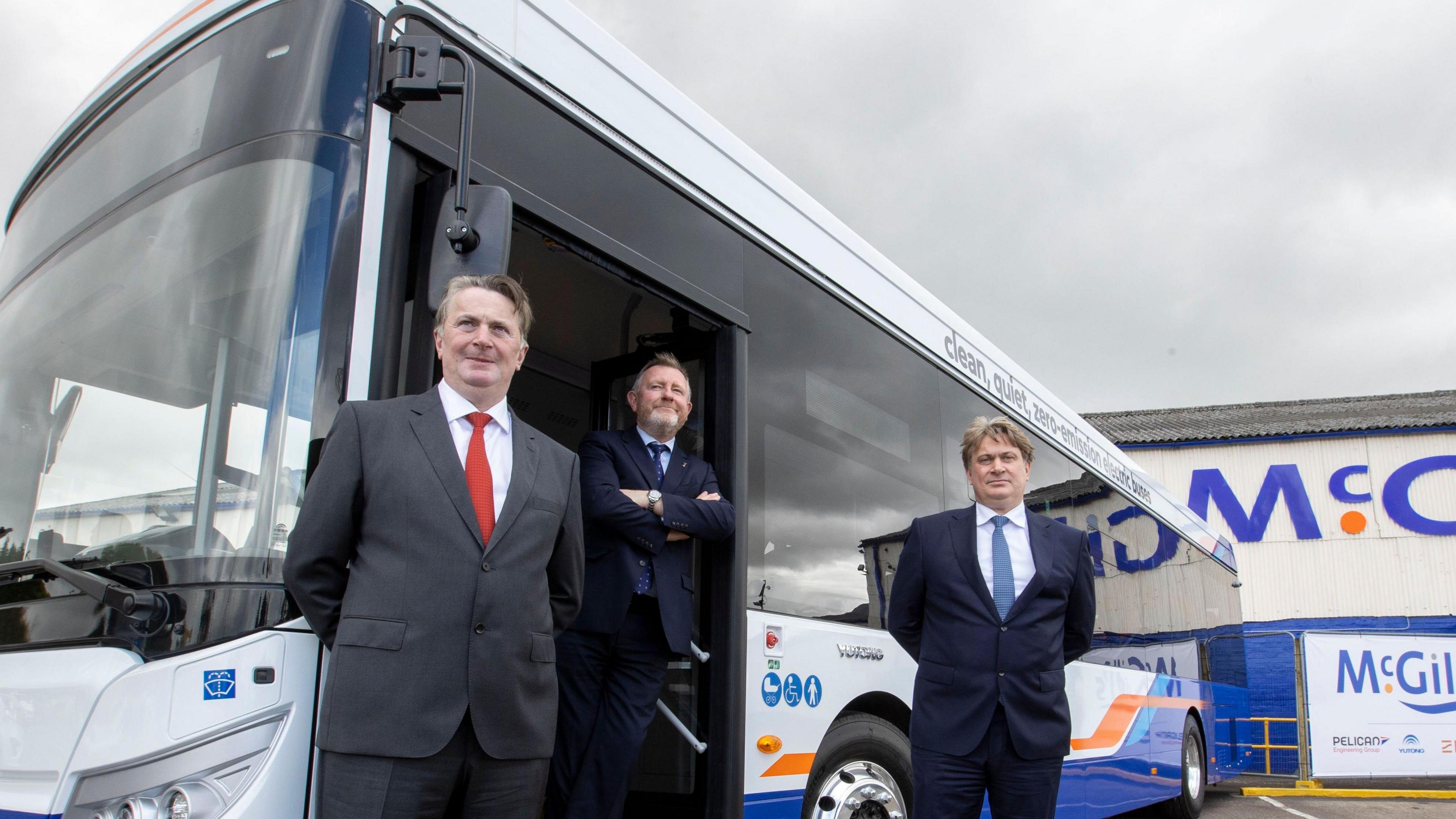 Three men, dressed in suits, standing next to a bus. It is parked in the car park of a large building.