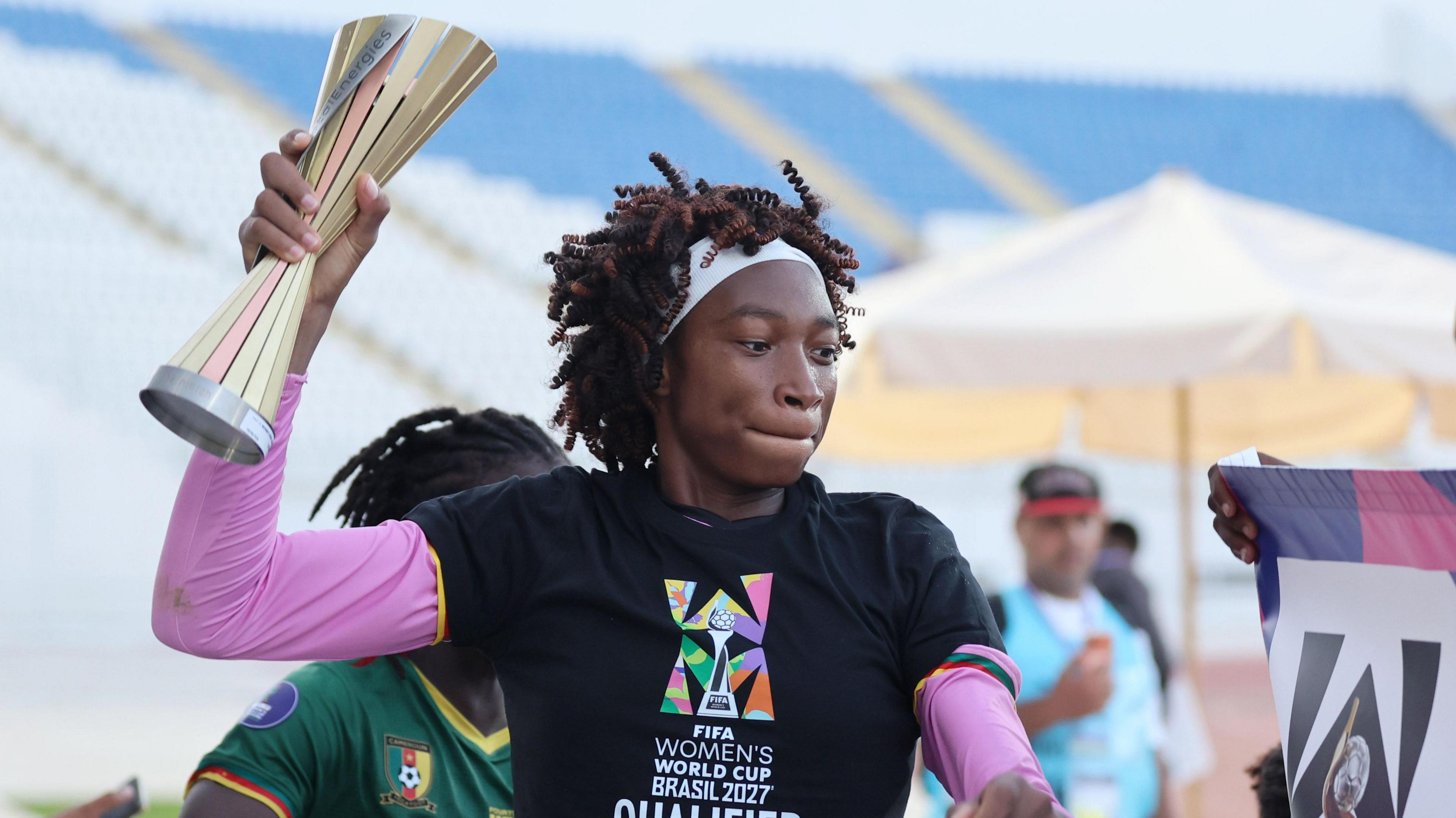 Cameroon goalkeeper Michaely Bihina, visible from stomach up, wears a black t-shirt bearing the message 'Fifa Women's World Cup Brasil 2027 qualified' over a long-sleeved pink top. She has a thick white headband holding back her hair as she holds up a player of the match trophy, which is disappearing out of the top of the image, with her right hand. A team-mate in a green Cameroon jersey is partially obscured behind her, while blurred in the background a man in a bright blue tabard watches on in front of the empty stand of a stadium