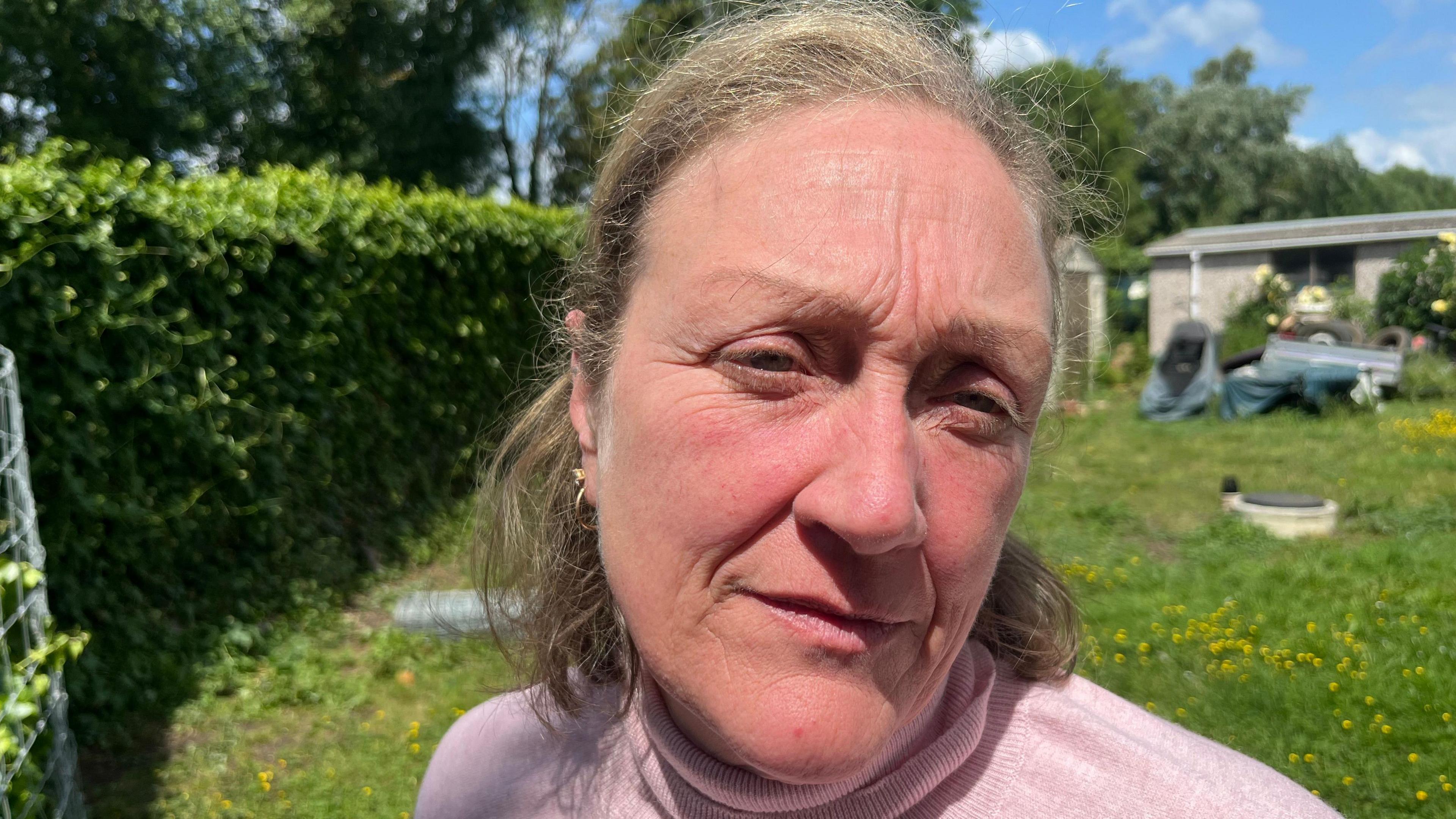 Becky wearing a pink knitted jumper- standing in her garden with green grass in the background looking into the camera.