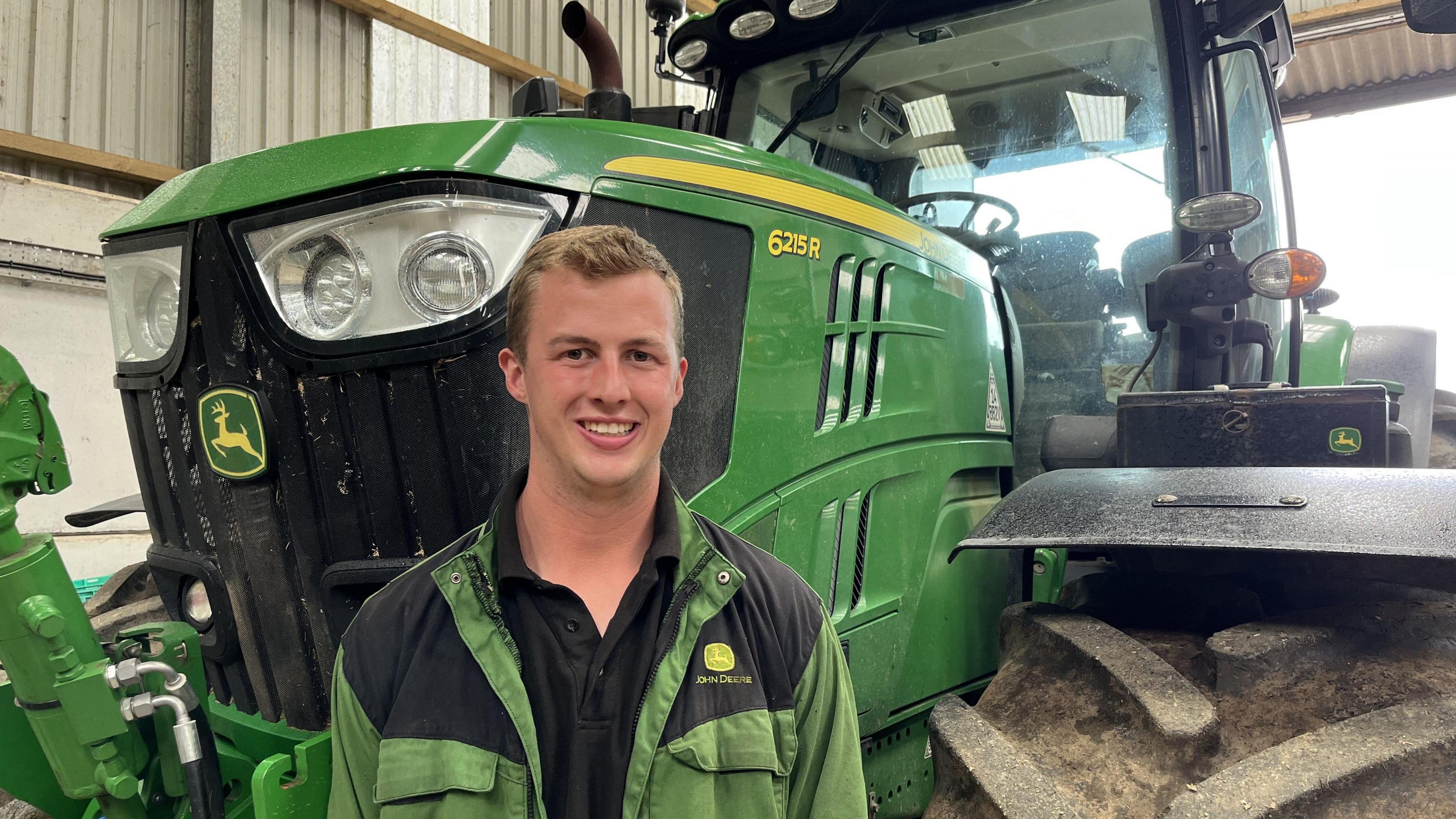 Harvey Jones is wearing green overalls. He is standing in front of a green tractor.