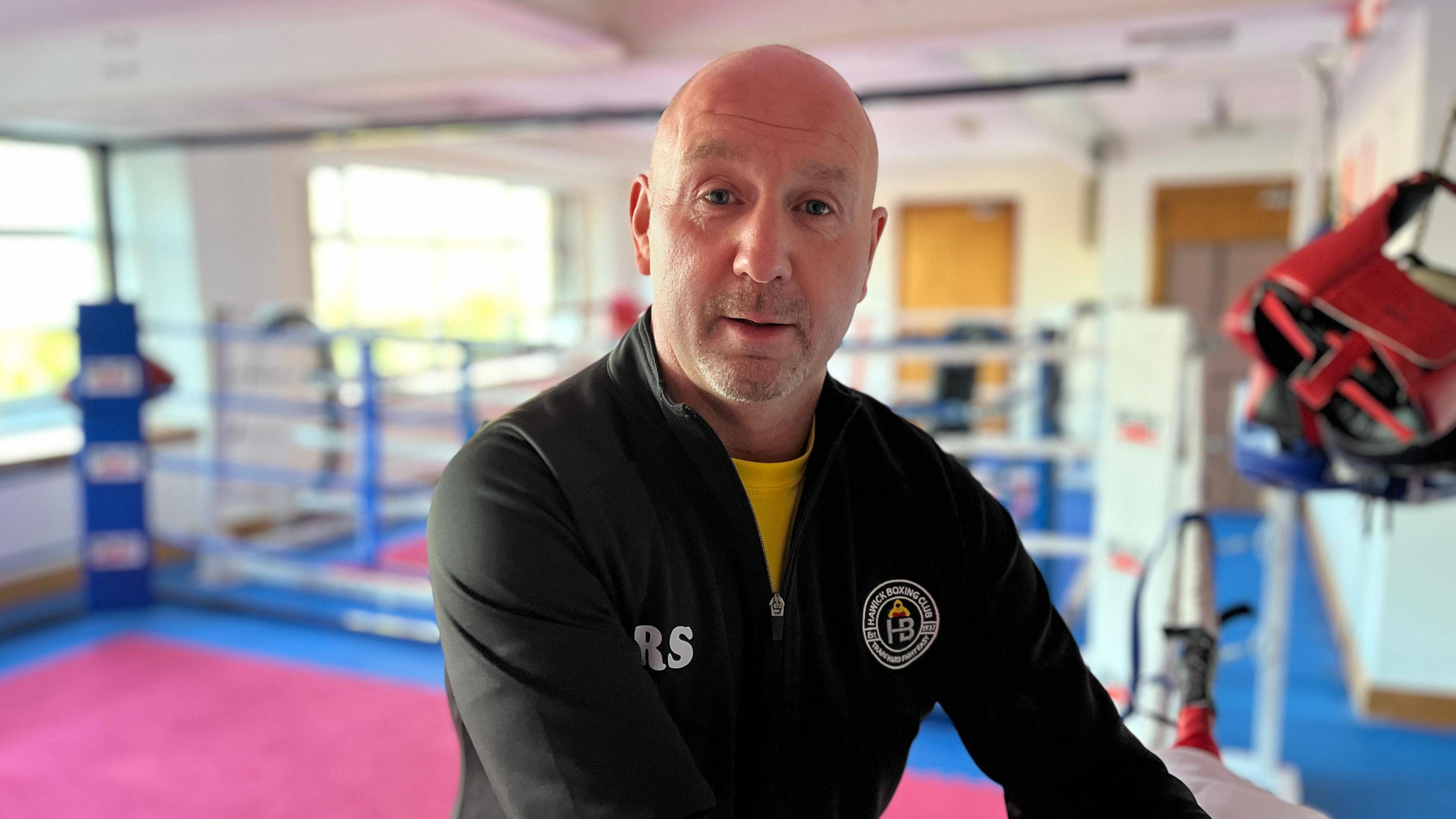 A bald-headed man, wearing a black top, look at the camera. Behind him is a boxing ring