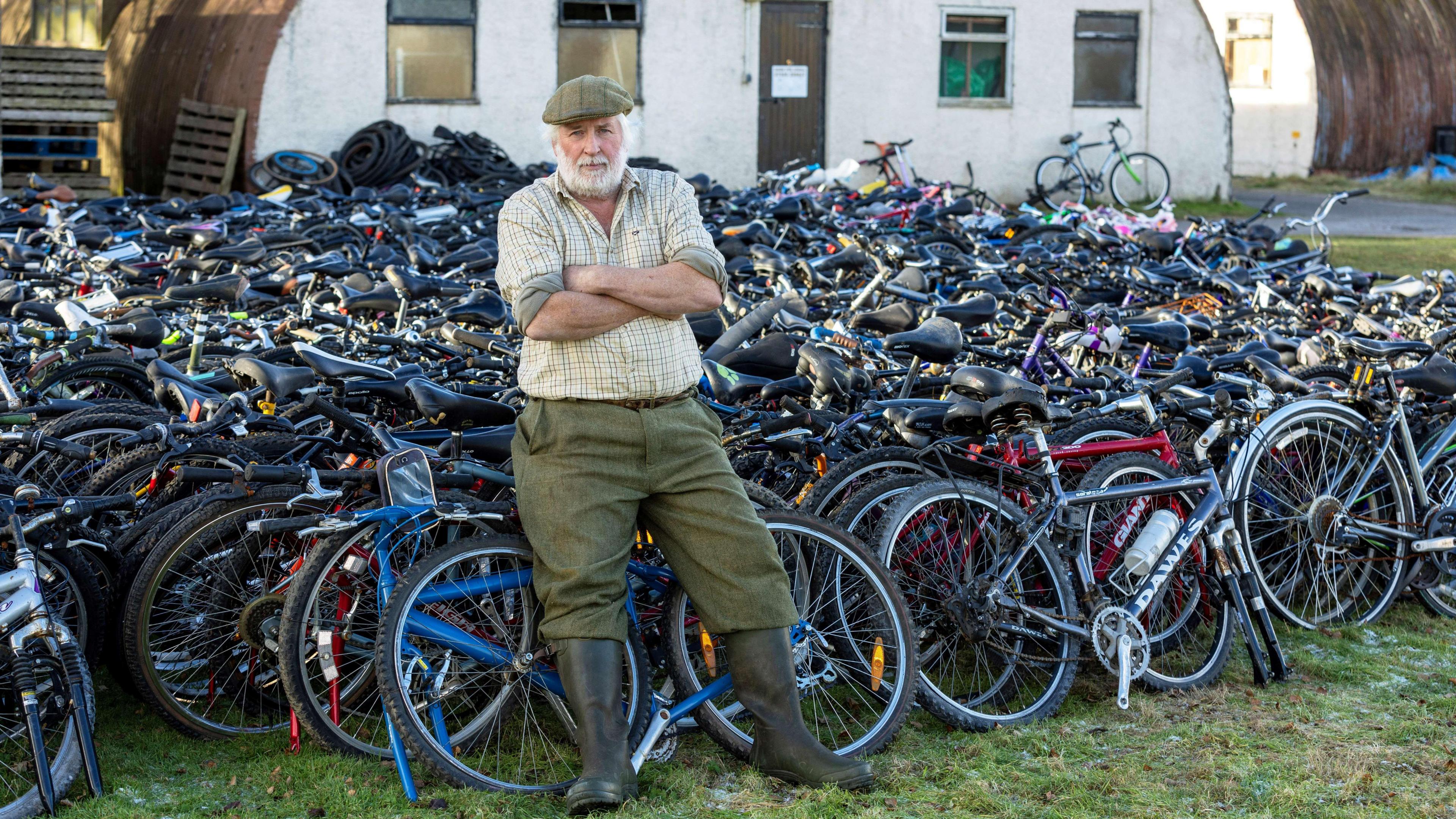 Nigel Carter leans against over a thousand bikes with his arms crossed looking at the camera. He has white hair and a beard, wearing a checked shirt and green trousers with black welly boots. 