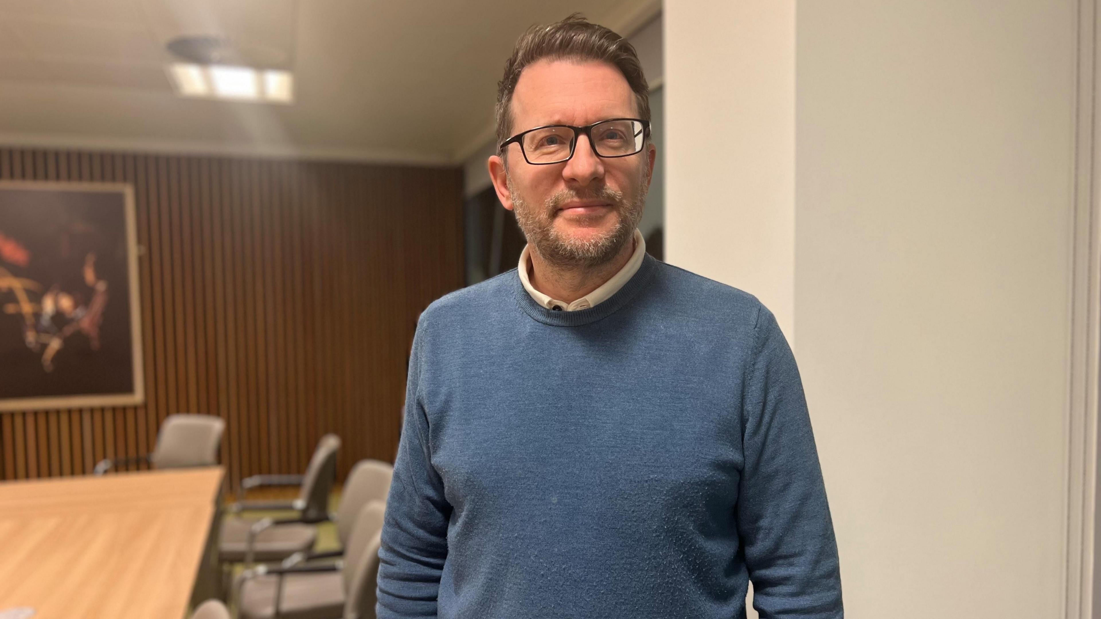 Man in blue jumper with short hair and glasses standing in room with a table and chairs 