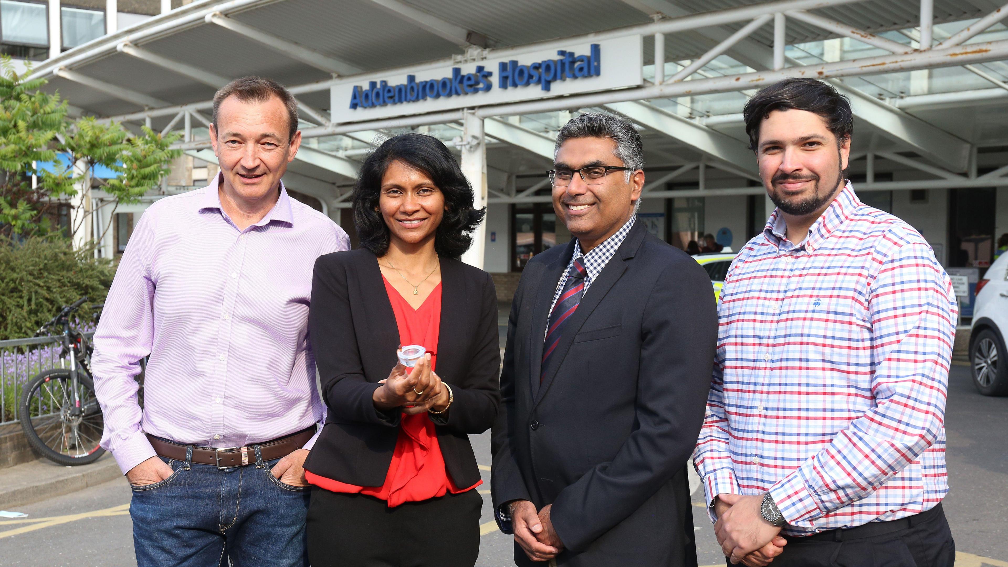 )Dr Alex Samoshkin, Professor Sohini Kar-Narayan, Affiliate Associate Professor Vikas Khanduja and Dr Jehangir Cama stand side by side outside Addenbrooke's Hospital. They are all smiling and looking directly at the camera. 