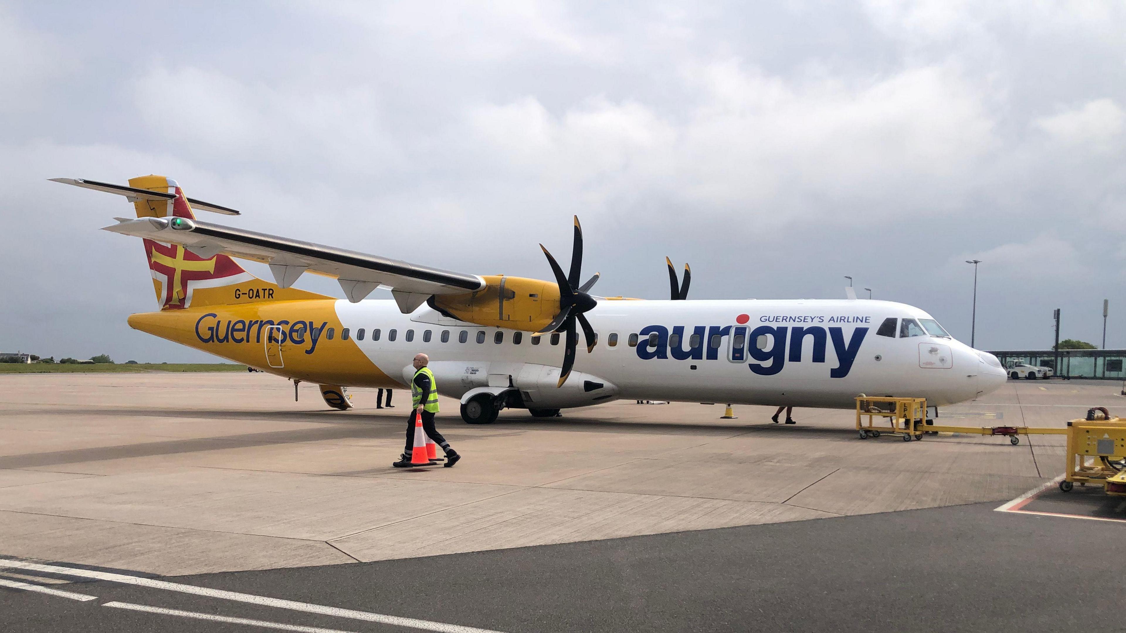 An Aurigny ATR plane on the runway at Guernsey Airport. It has a white and yellow livery and propellers.