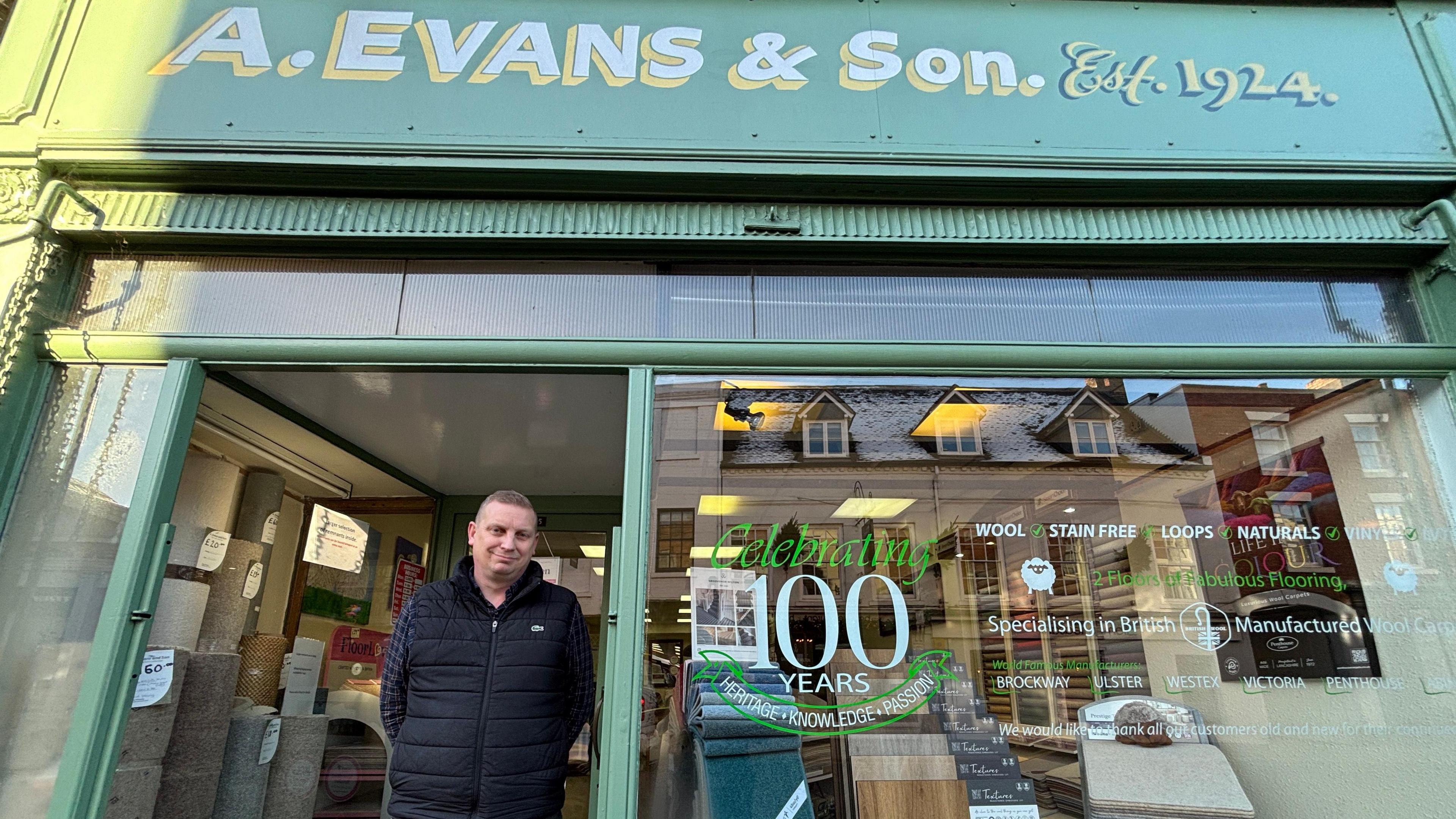 Mr Edwards has short brown hair and is wearing a black gilet over a dark blue shirt. He is standing in the doorway of the shop. The shop's exterior is green with a painted logo that reads "A Evans and Son, established 1924". There is a sticker on the shop window which says "Celebrating 100 years"