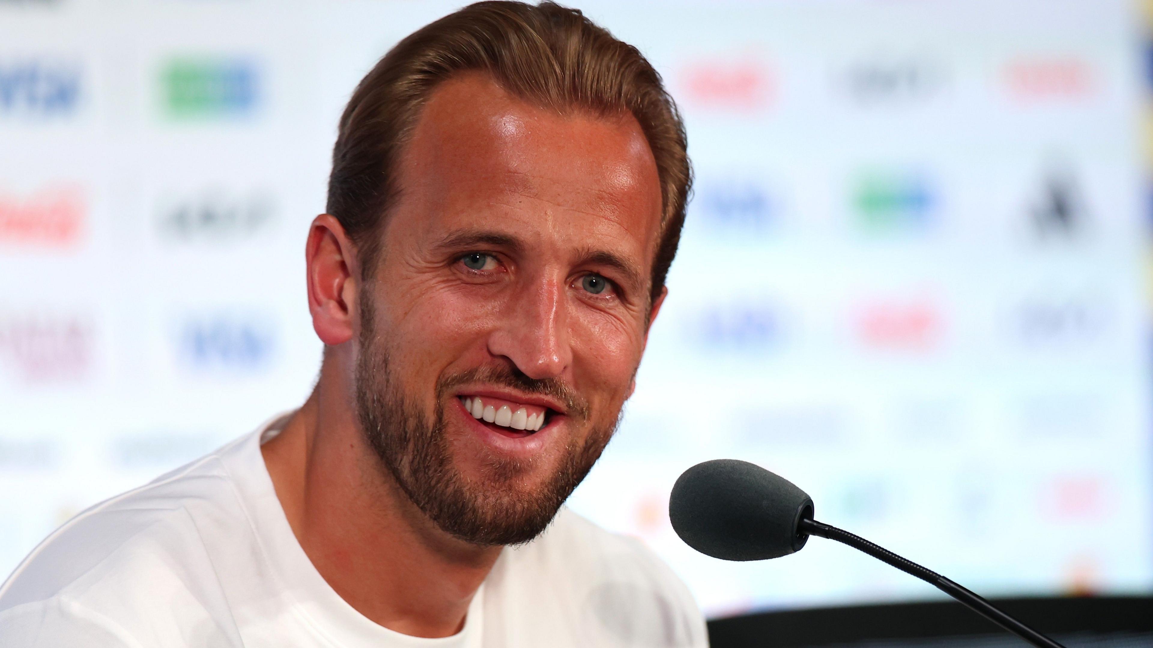 Harry Kane sits by a microphone at a press conference, looking out and smiling