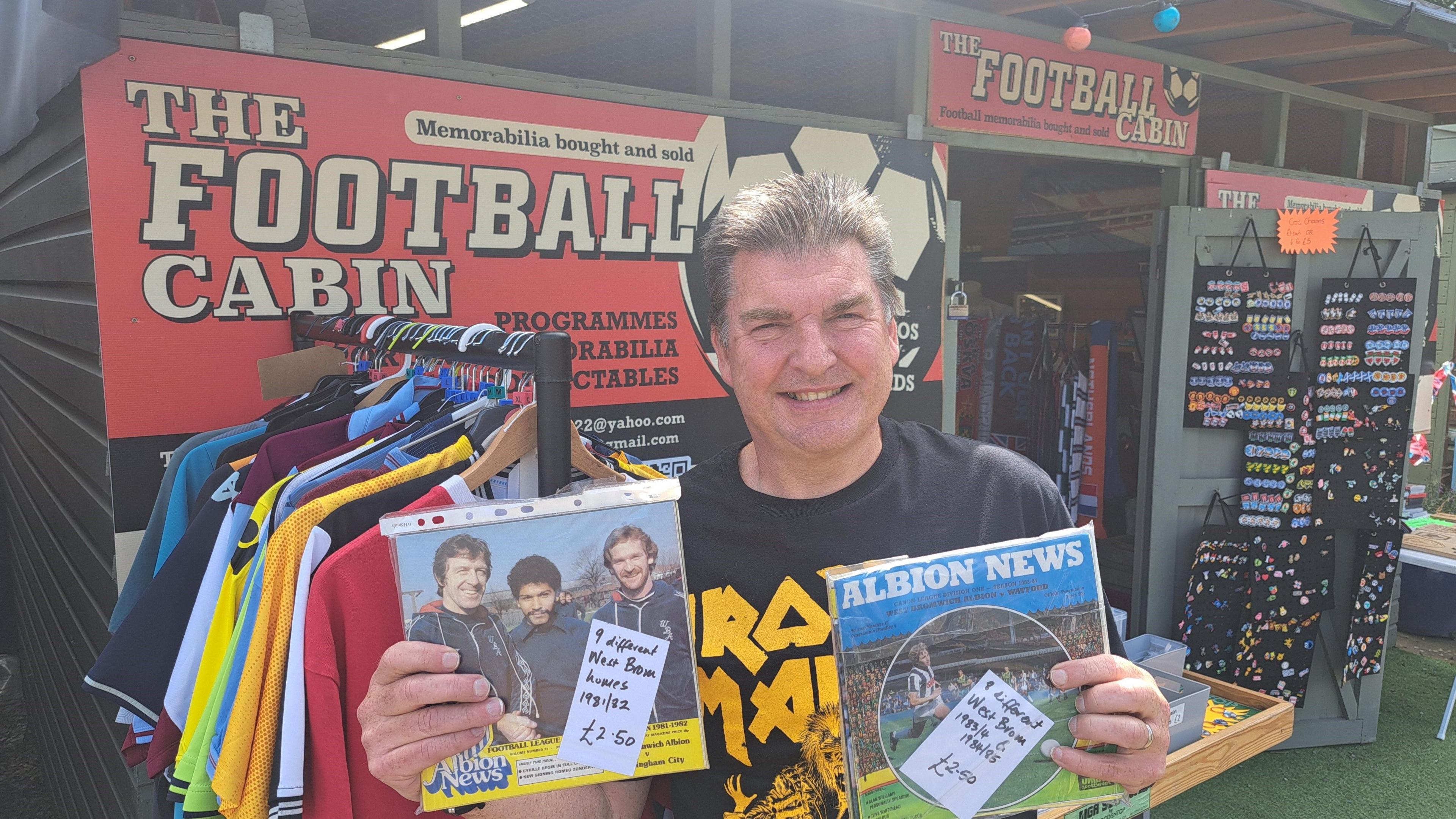 A man holding two football programmes is standing outside the premises of The Football Cabin. There are two signs with its name and football shirts are hung up on the rail outside.