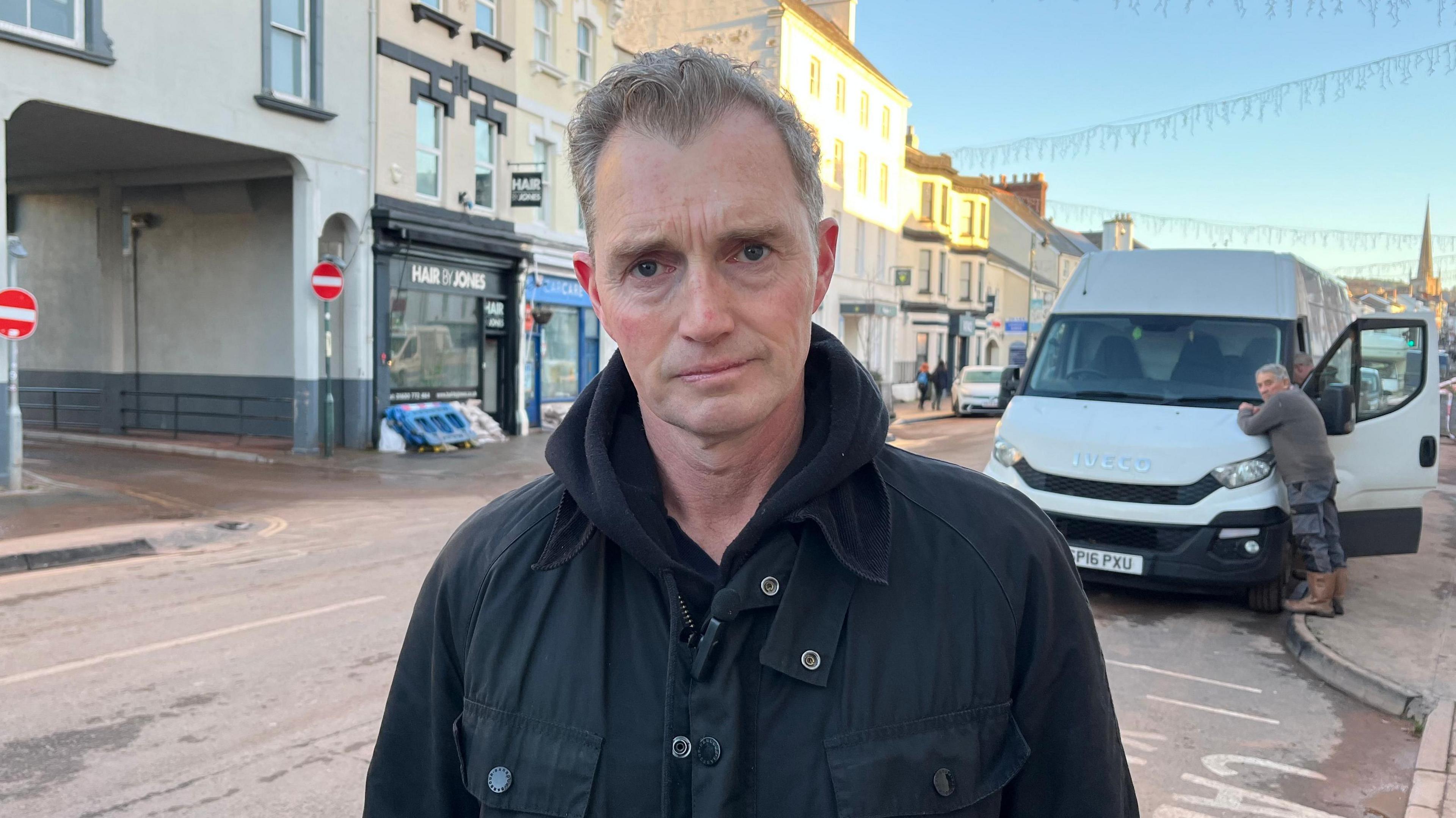Davies Davies is wearing a black coat and is standing in on a pavement of a high street. Behind him are flats and shops, and a large white van with a person leaning on its bonnet.