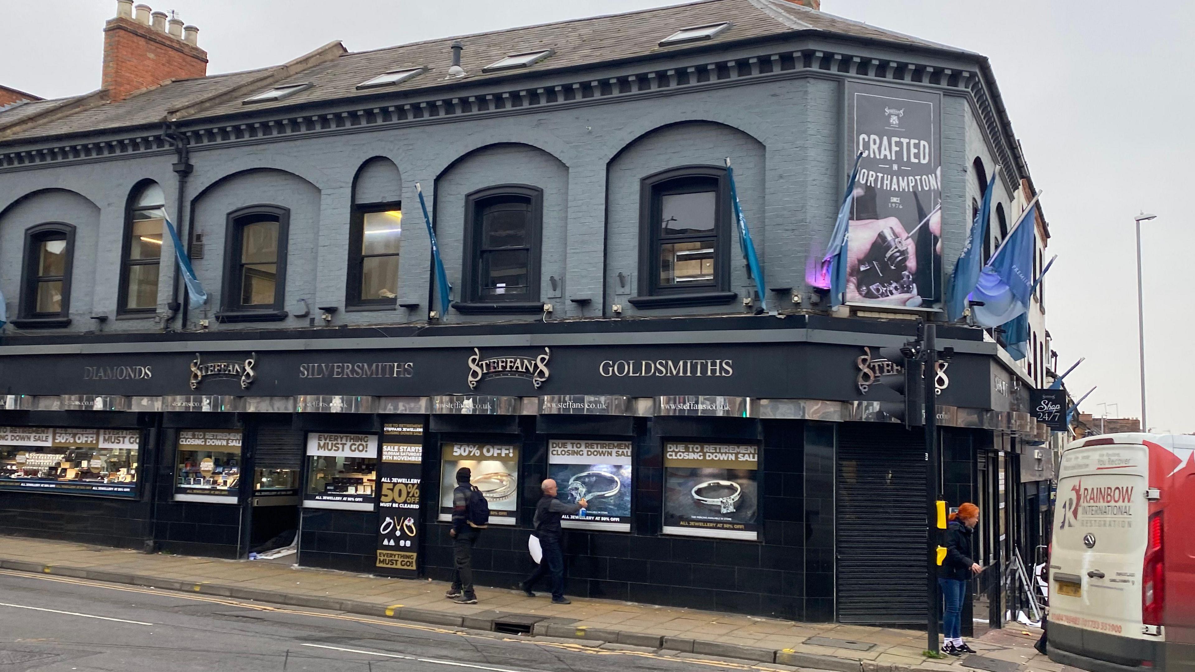 A photograph of Stefans Jewellers from the street. A black and dark grey building displaying jewellery with signs saying it is closing down and customers can receive 50% off.  There are three people outside of it, two of which are looking into the windows. 