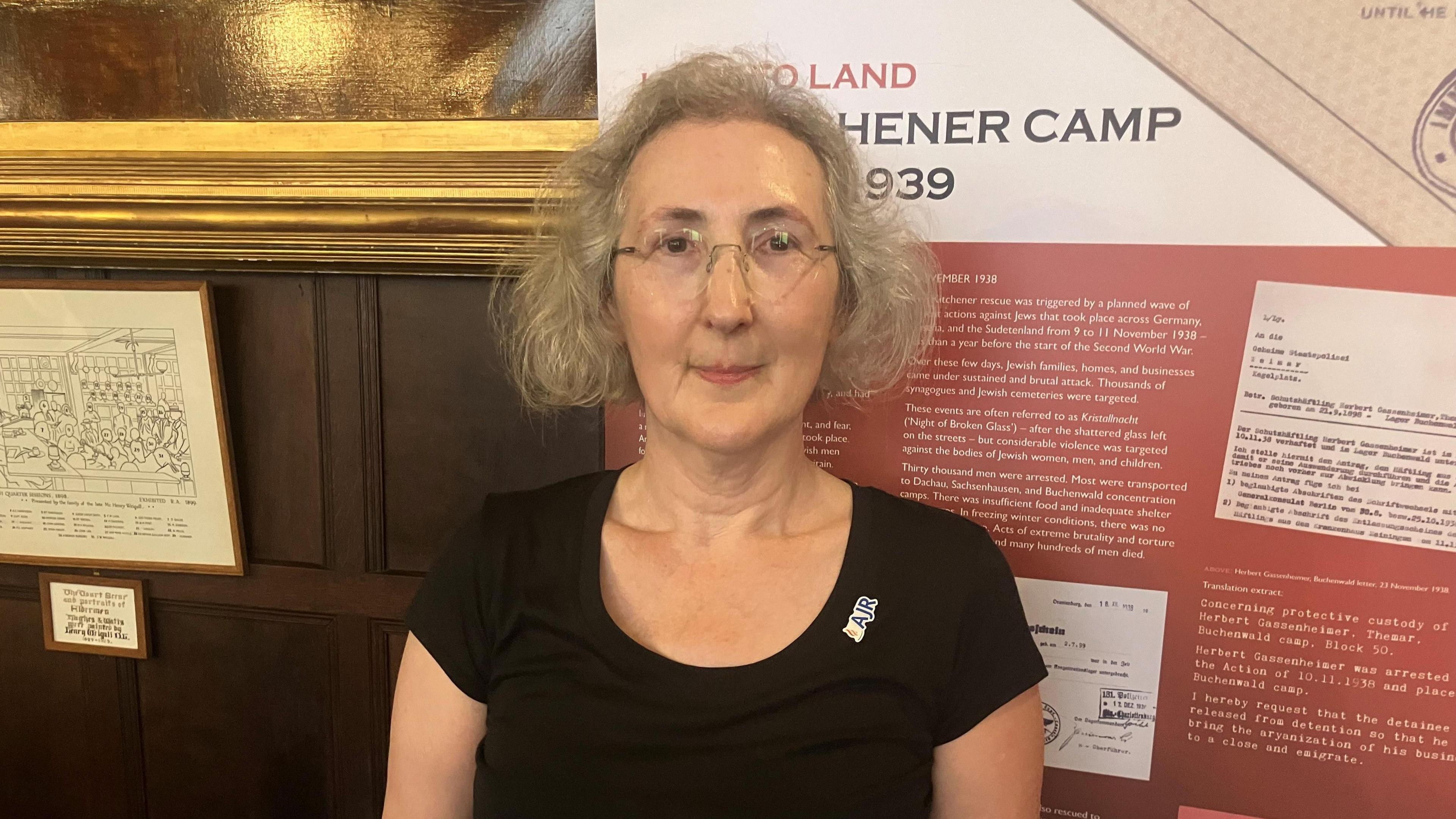 Clare Weissenberg - founder of the Kitchener Camp Project. She has grey hair, wears glasses and a black top. Behind her are items from a museum exhibition on the subject