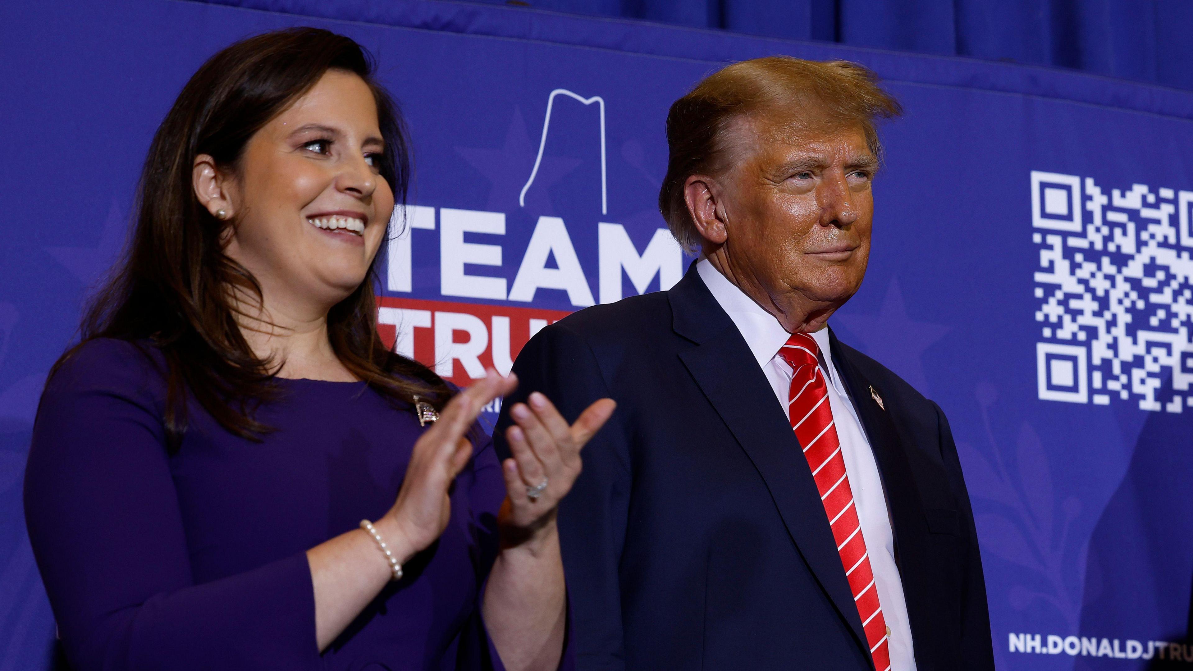 Elise Stefanik wears a blue dress and claps her hands. She is standing on a stage next to Donald Trump, who wears a dark suit and red tie.