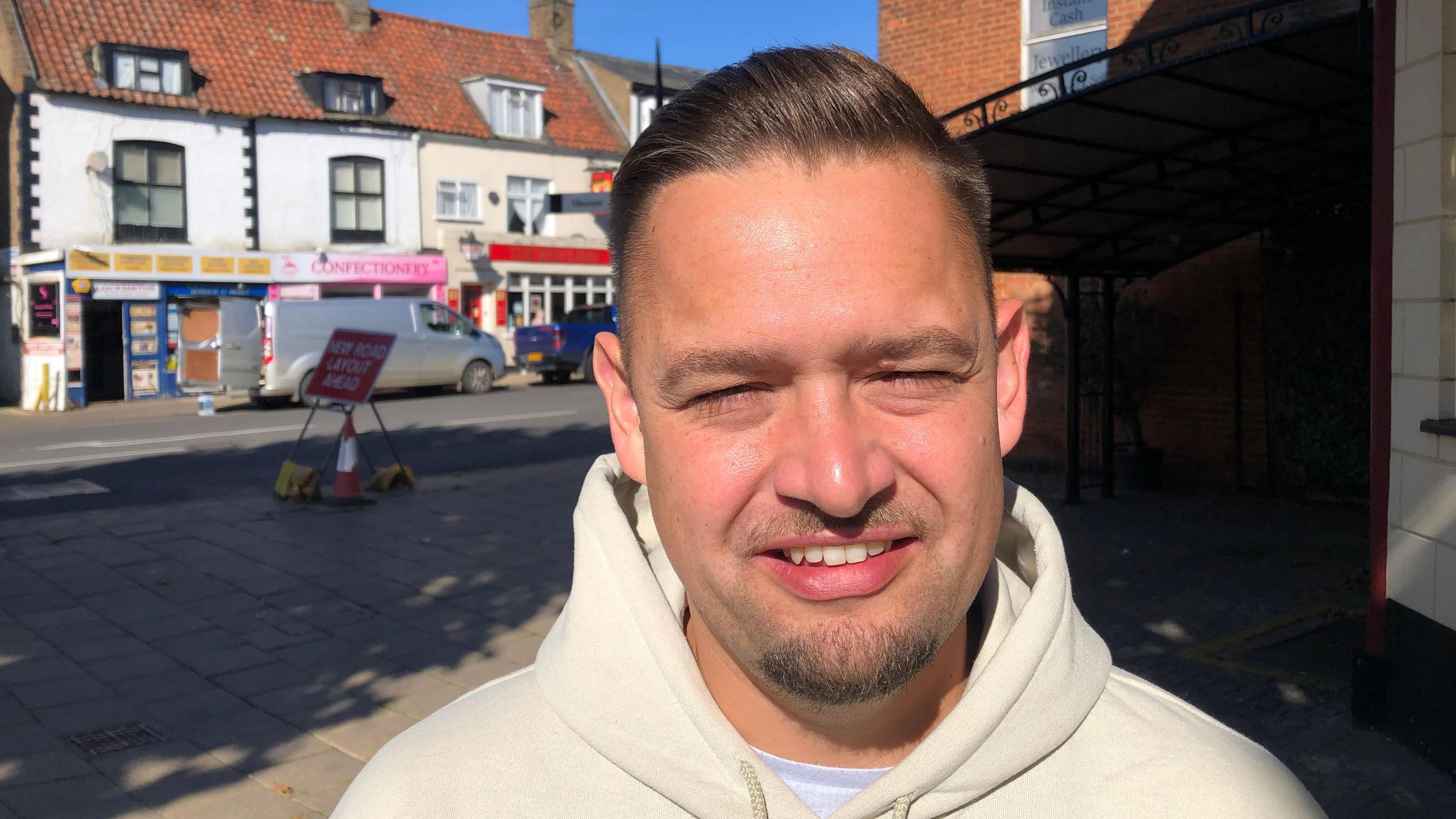 Daniel Gray smiles at the camera as he squints as he stands directly in the sun. He is wearing a white hoodie sweatshirt and is photographed on a high street with shops in the background. 