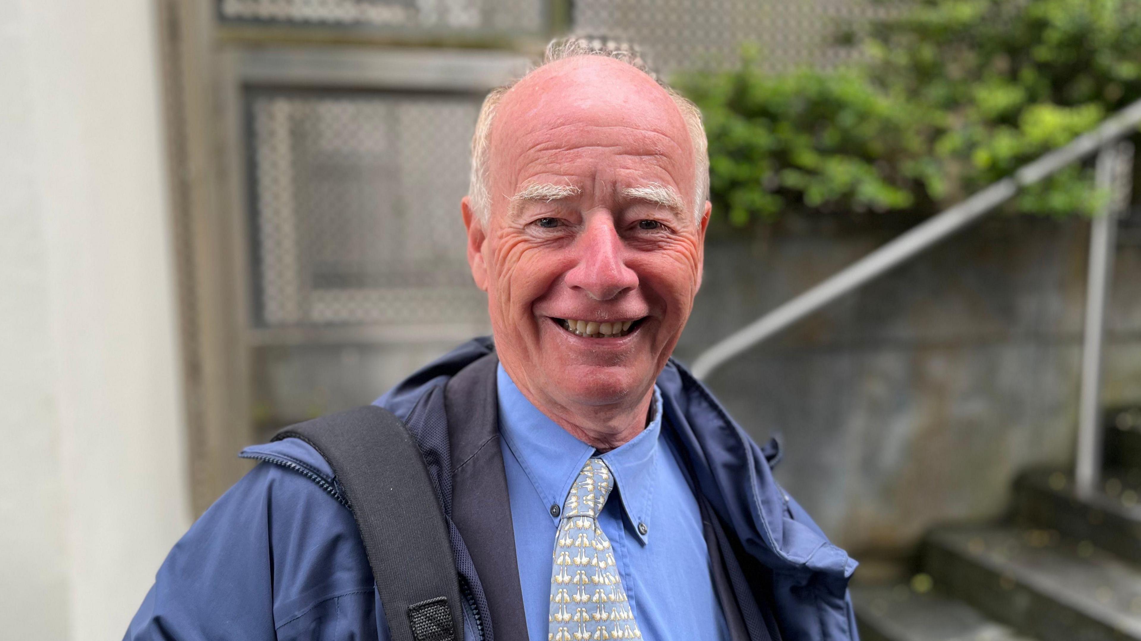 Deputy George Oswald - A man with a bald top of his head, with white hair on the sides. He's wearing a blue raincoat, blue shirt with a blue and white patterned tie. On his right shoulder is the strap of a backpack.