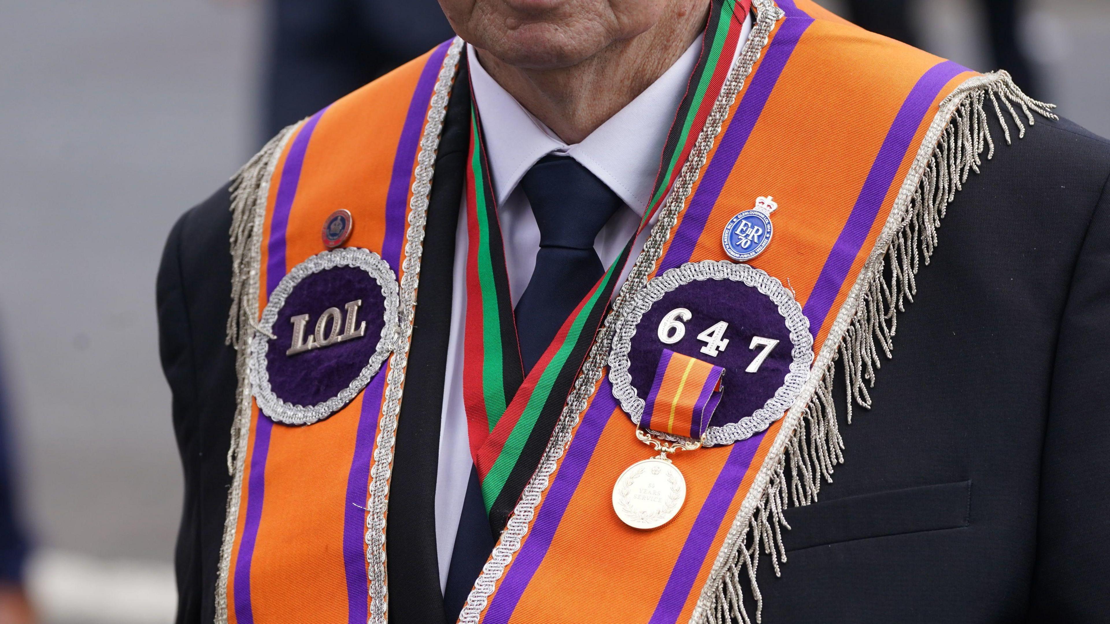 The shoulders of a man in a dark suit who is wearing an orange sash with the emblame of the Loyal Orange Lodge, and the lodge number 647. He also has a lapel badge marking the 70th jubilee of the late Queen Elizabeth the second. 