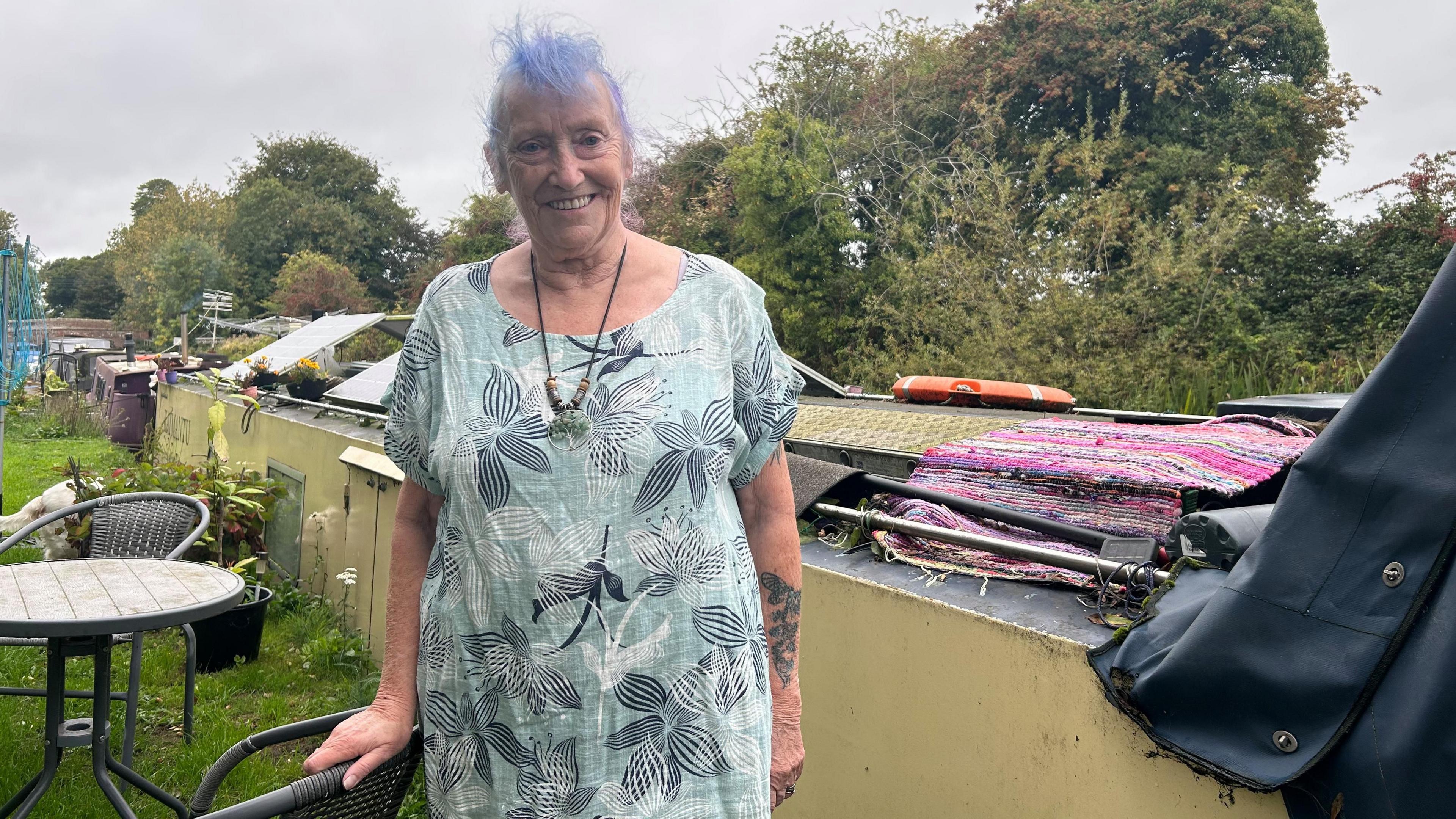 Lyndsey stands smiling outside her canal boat wearing a pale green dress with large white and black flowers on it. She has light blue hair and wears a handmade necklace which has brown beads and a tree dangling from it. Her boat in the background is pale yellow with two solar panels on top. To her left on the grass is a black woven garden table and chair set. 