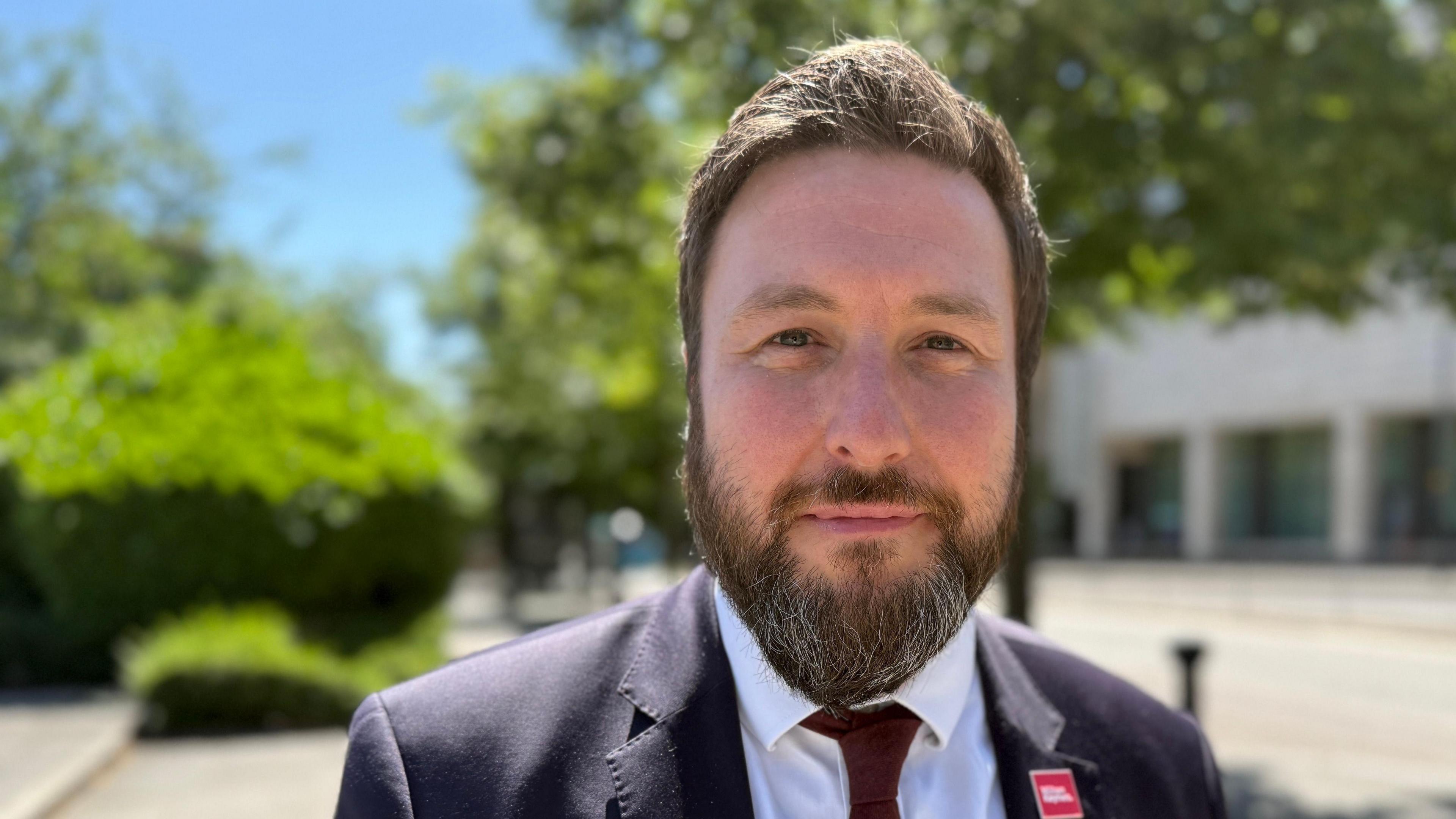 A head and shoulders image of Pete Marland. He is wearing a dark blazer over a white shirt with a dark red tie. He is looking into the camera and blurred trees are in the background. 