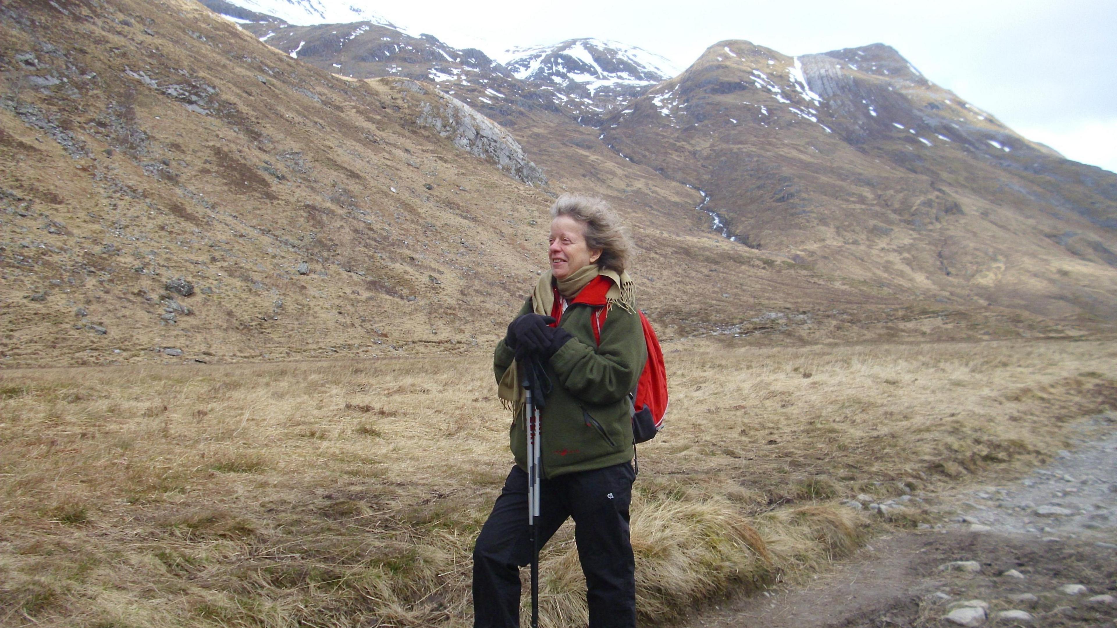 Alison wearing a green jacket, black trousers, red backpack and holding walking sticks in front of a mountain