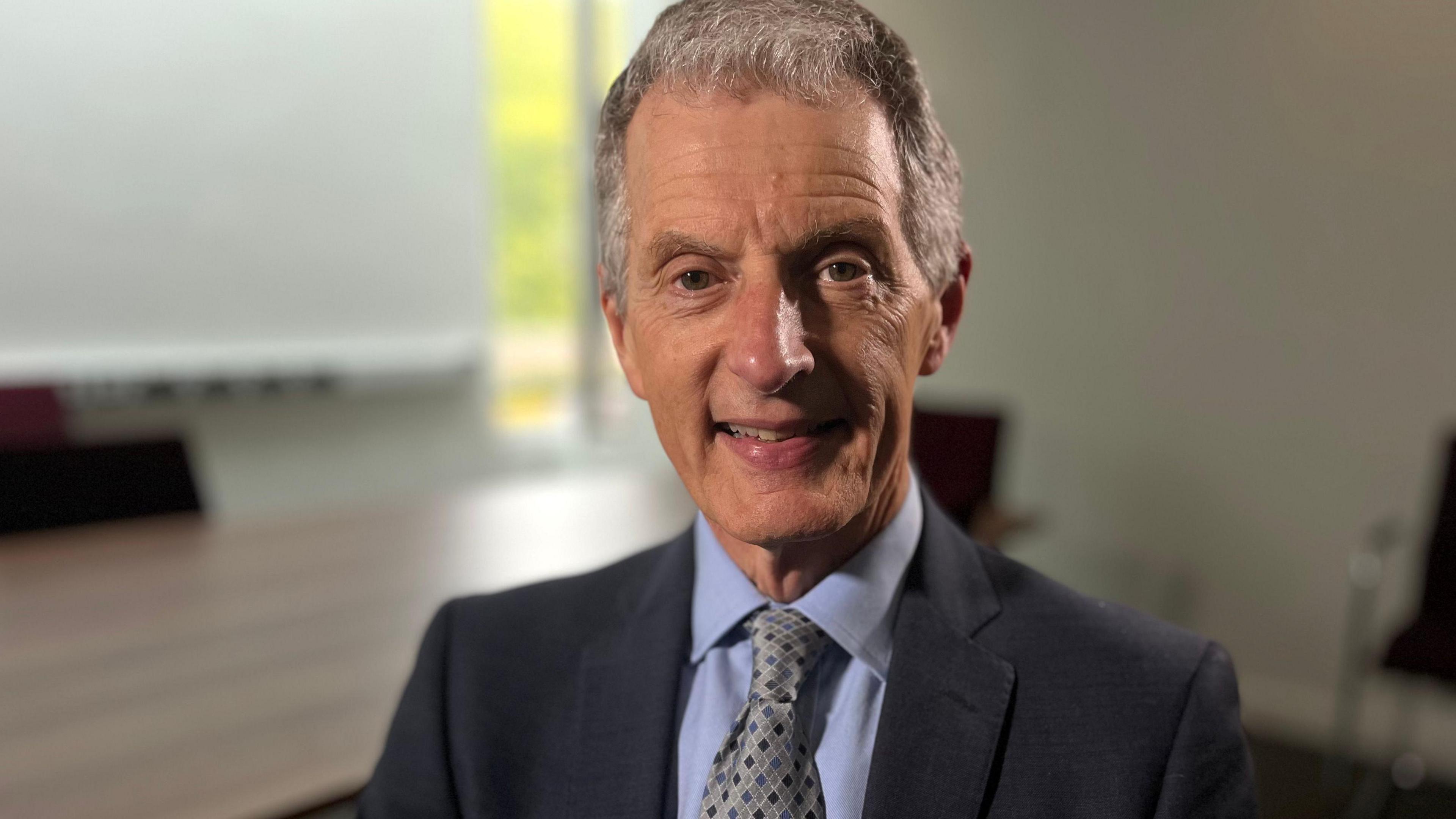 David Maguire with short cropped grey hair, drawn forward slightly on his forehead. He is wearing a blue suit jacket, a light blue shirt and blue tie and is smiling at the camera with a blurred out office table behind him