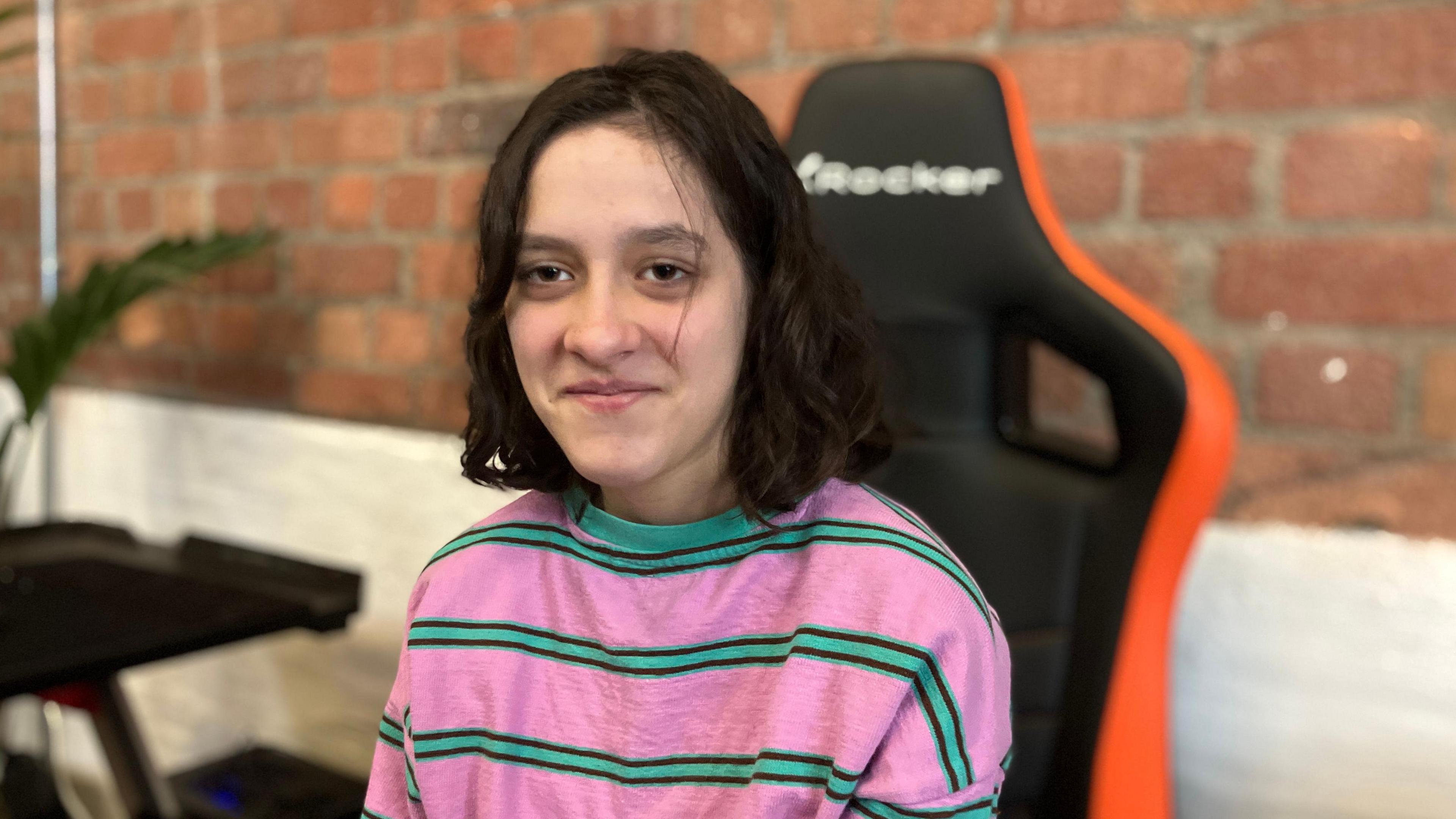 Student Iris, wearing a bright pink jumper, with horizontal green and black stripes. She is sitting in a black gaming chair with an orange outline against a brick wall and is smiling.