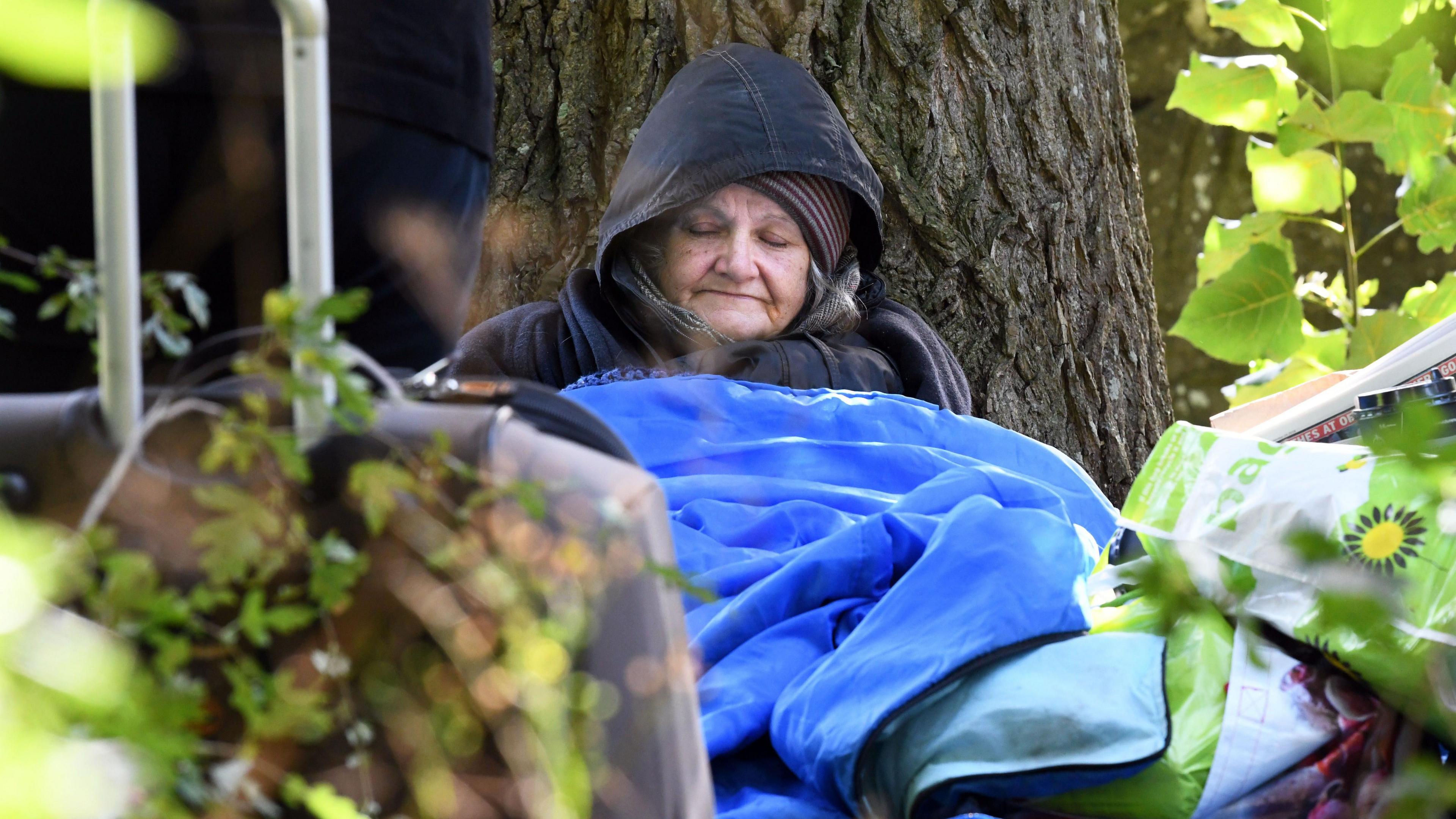 Ann Hillman in a blue sleeping back sits against a tree sleeping. She is wearing a dark coat with the hood up, and a stripped wooly hat. 