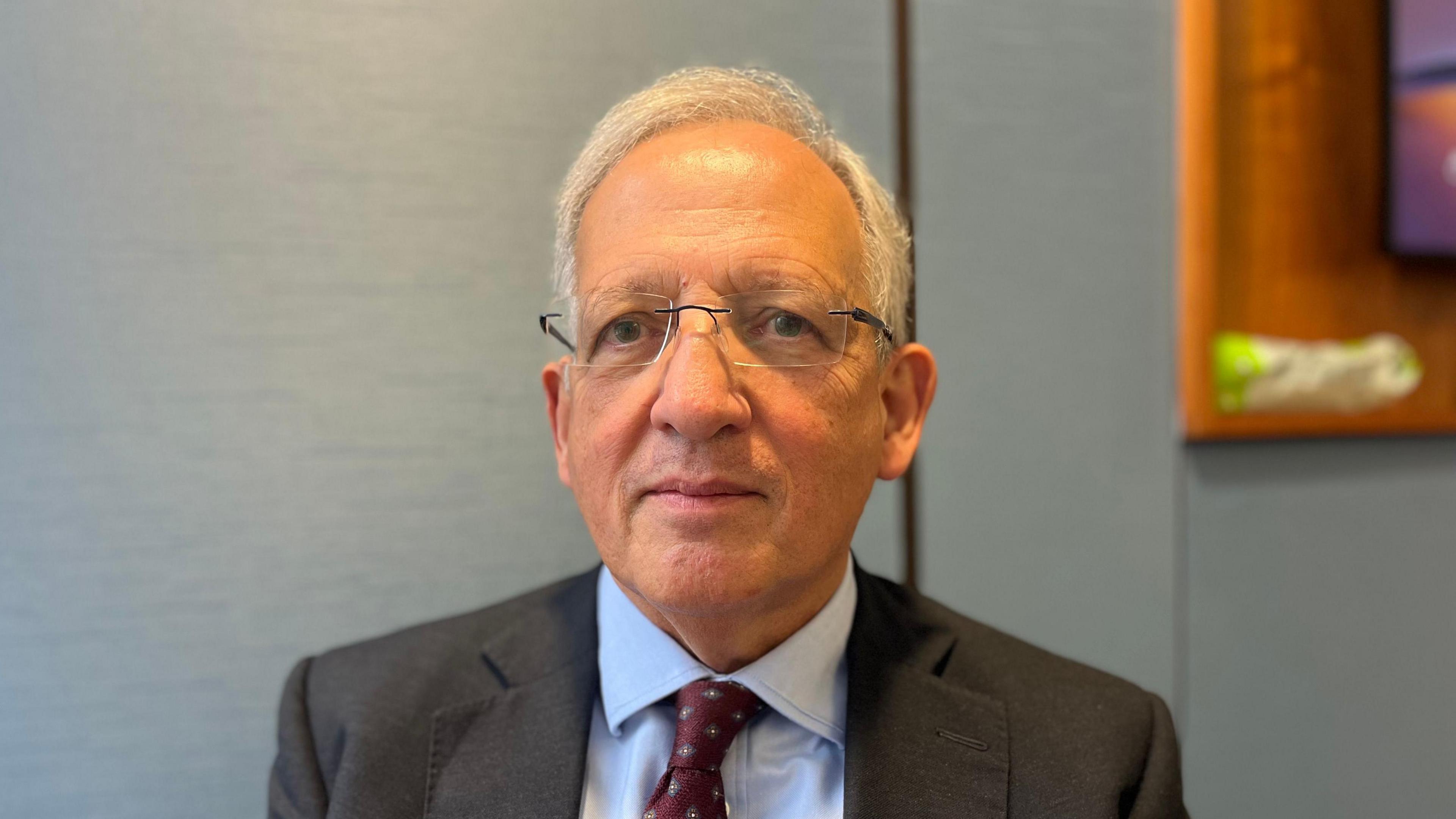 Jon Cunliffe is a white man with grey hair and glasses. He is staring into the camera. He is wearing a dark grey suit with a pale blue shirt and burgundy tie. He is in focus with a blurred blue background behind him.