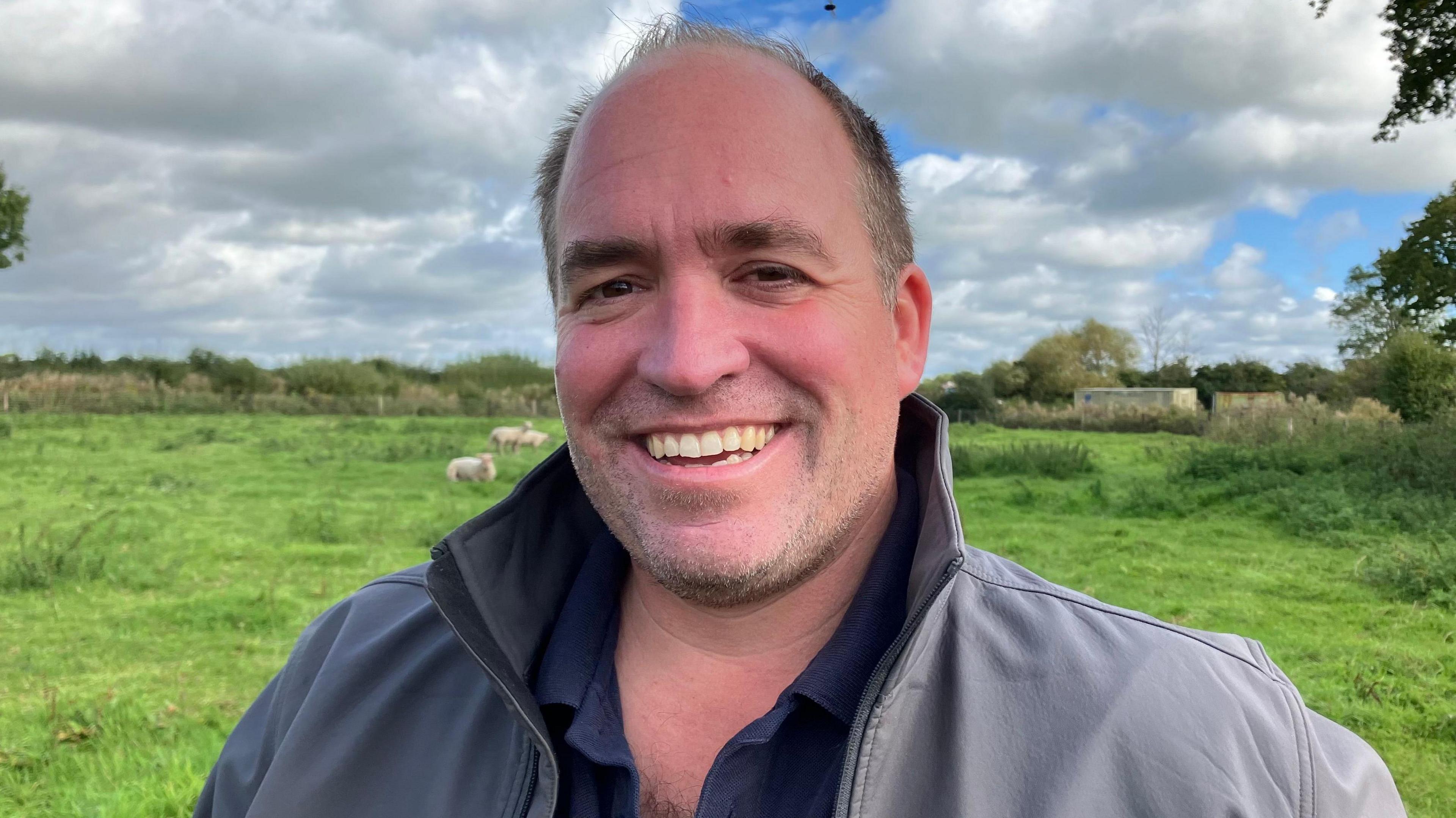 Farmer Ed Simmonds smiling at the camera standing in a field of sheep