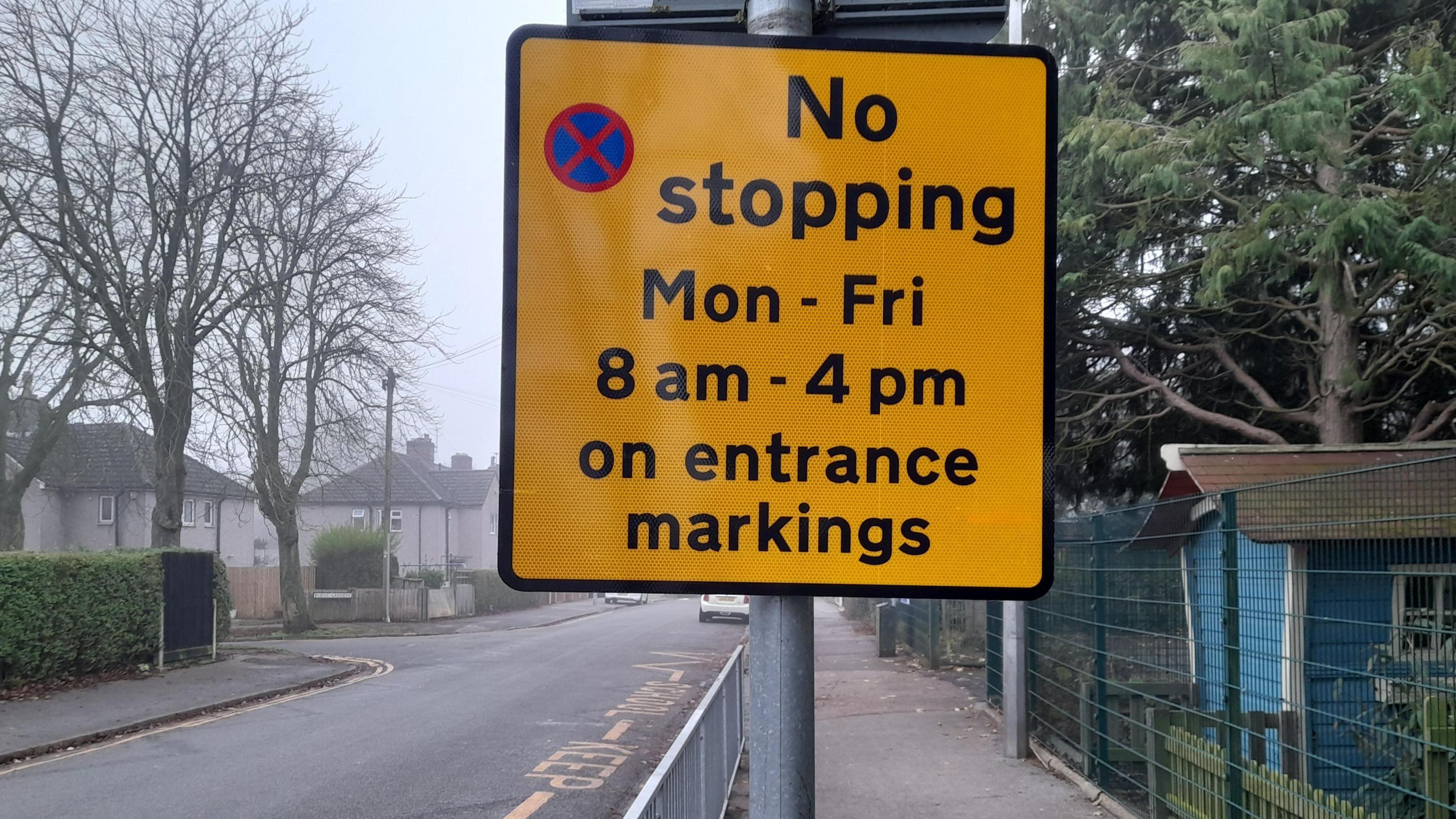 A yellow sign showing parking restrictions outside a school, with keep clear road markings on the road