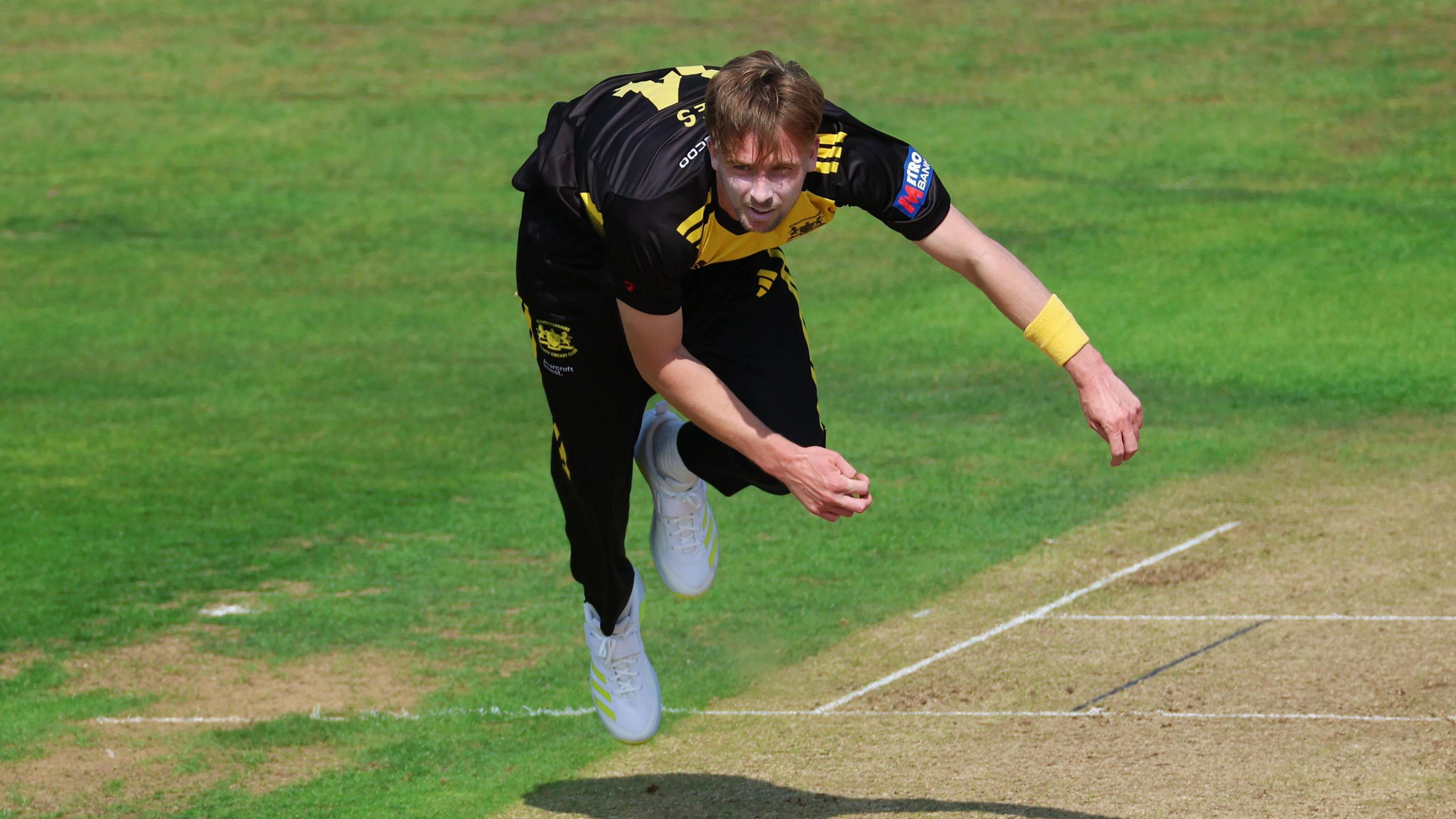 Gloucestershire bowler Craig Miles is airbone after delivering the ball during the 50-over match against Glamorgan at Bristol. He has short blonde hair and is wearing a black kit with yellow markings and white sports shoes

