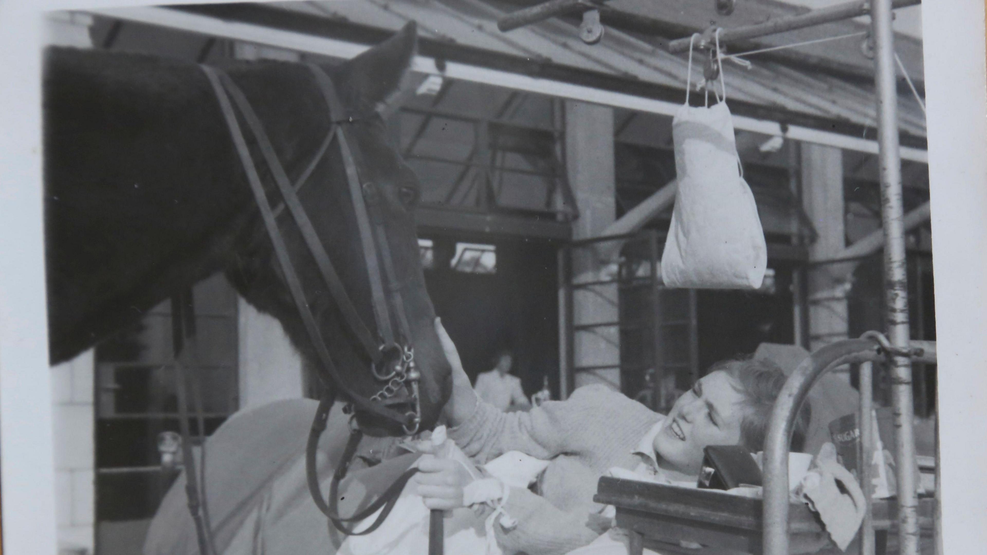 A black and white photo shows a young women in a hospital bed with a horse next to her. She is stroking the horses head and smiling. 