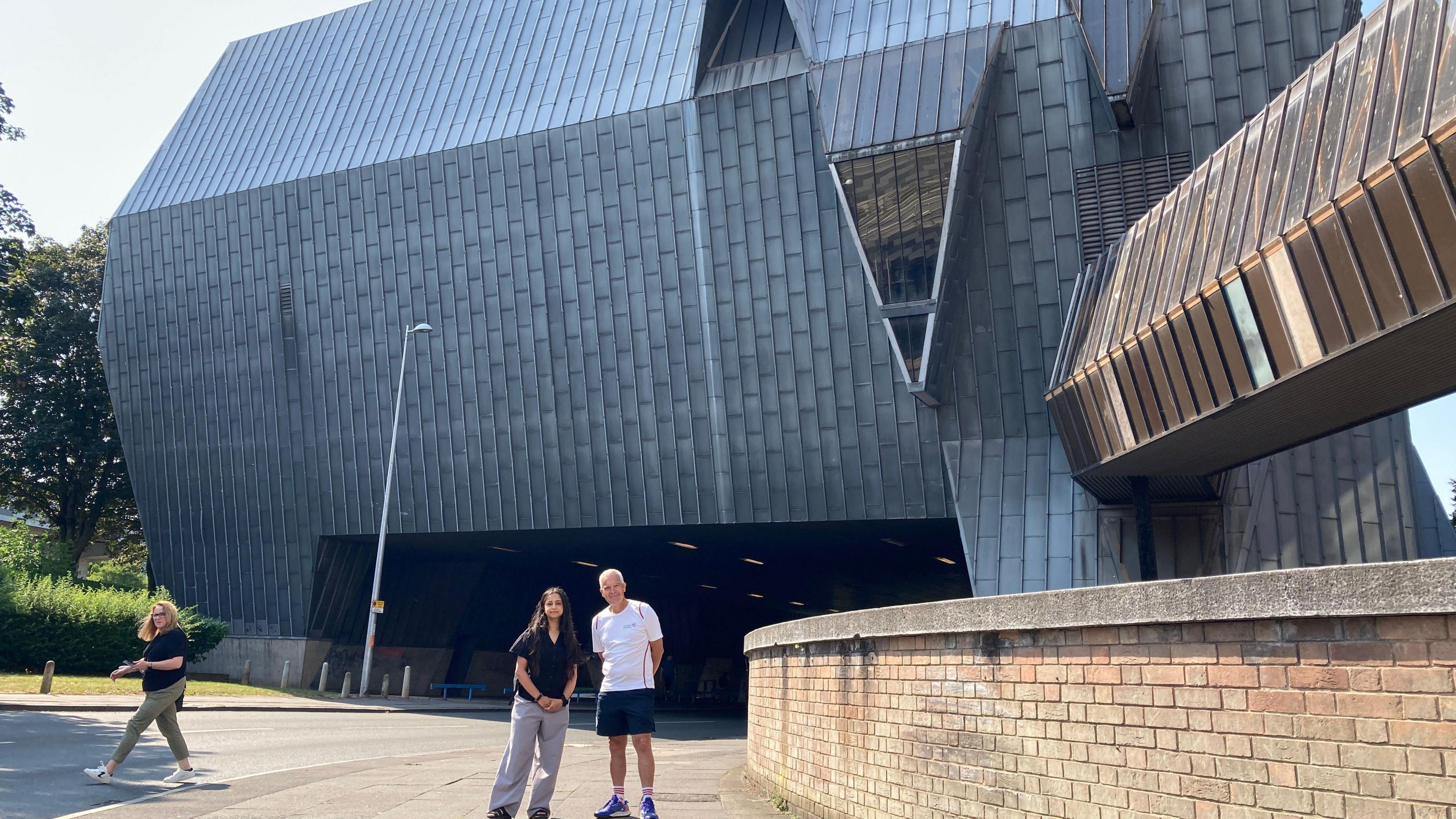 Two people are standing in front of a large, metal elephant-shaped building with a road running underneath it.