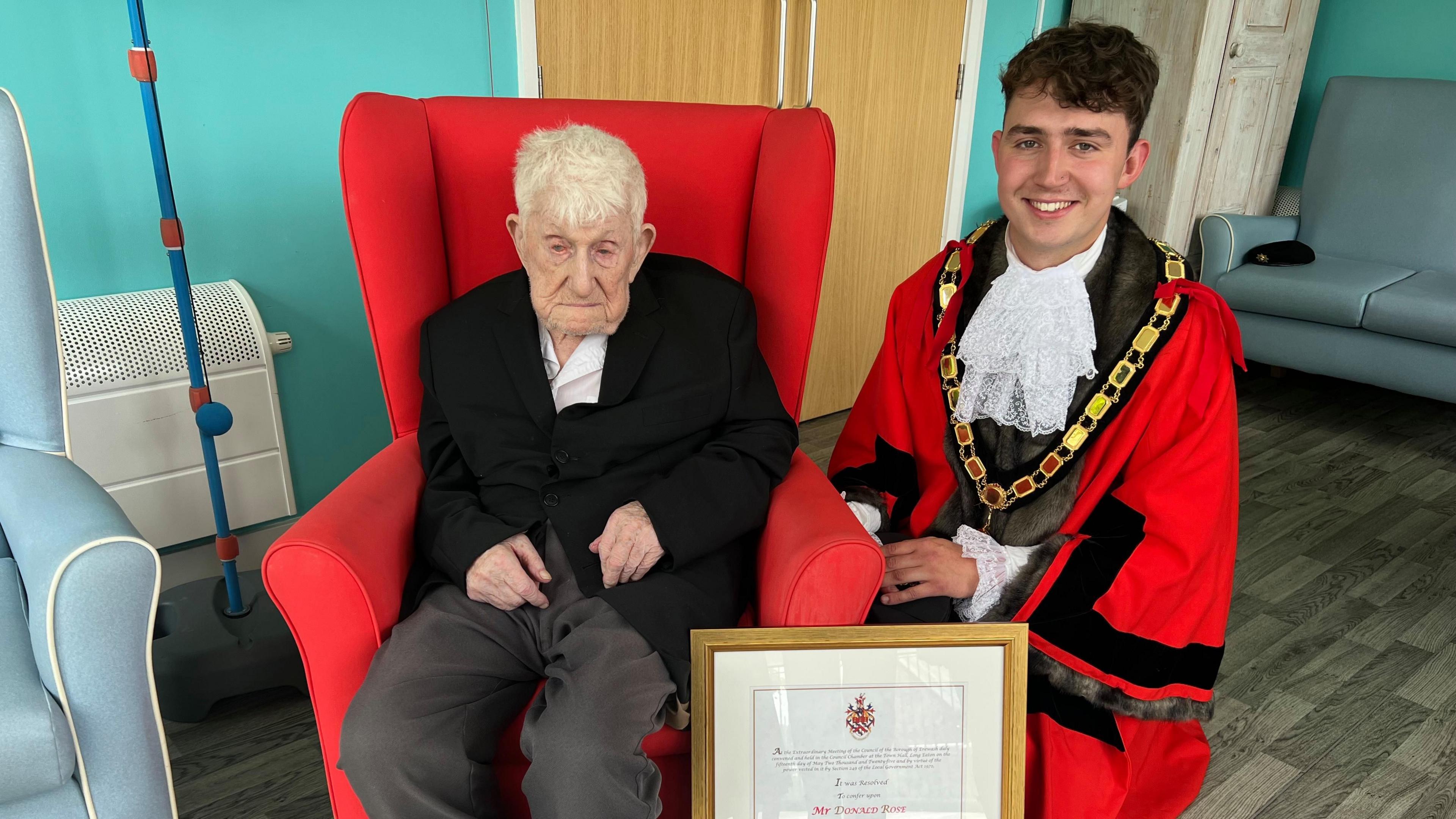 Old man in black blazer and grey suit trousers sat in red arm chair , young man with brown hair in ceremonial mayoral robes knelt beside him, wooden certificate leant on chair